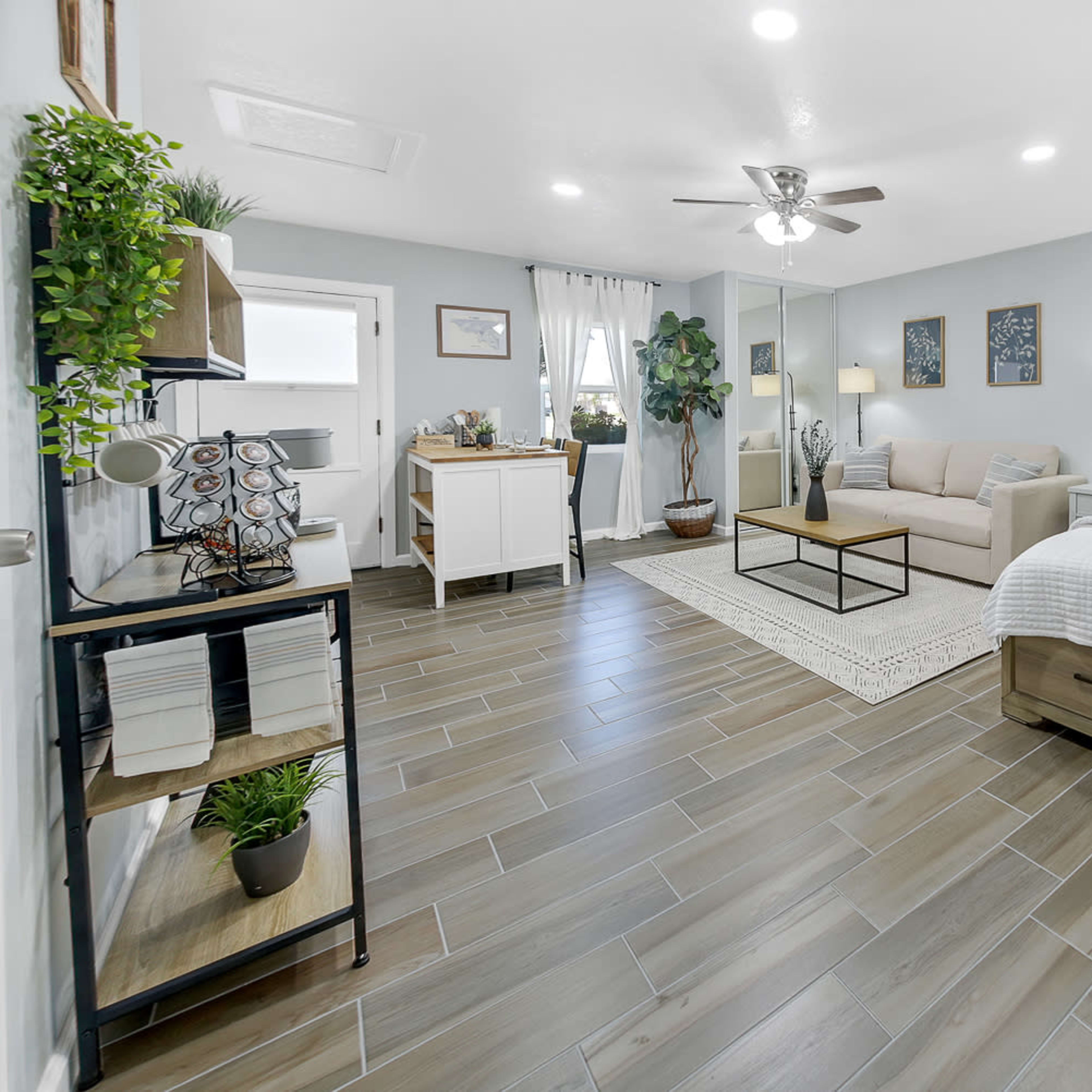 A modern studio room featuring a bed, a sofa, and a small dining area, with light-colored walls and wooden tile flooring.