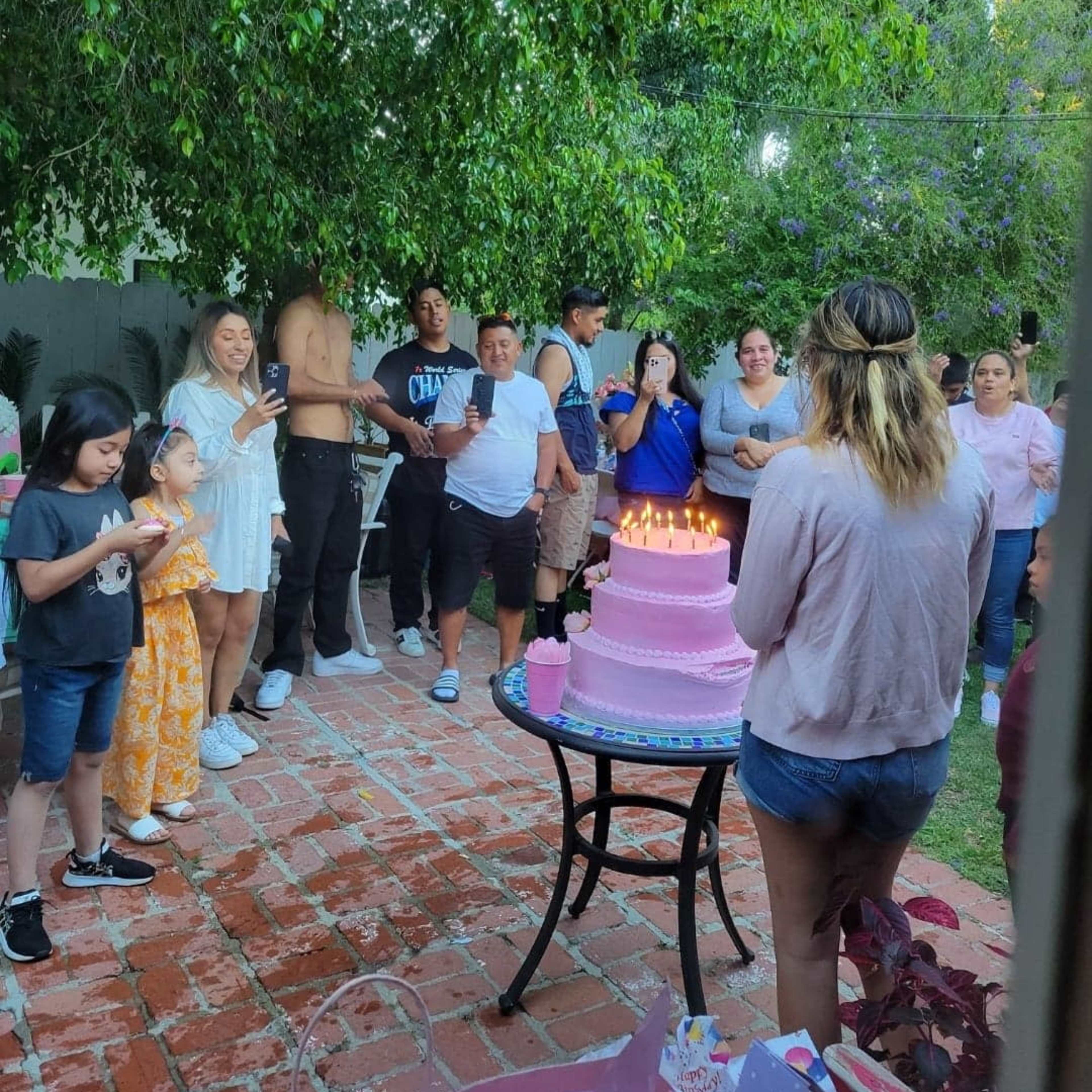 A group of people gathers in a backyard to celebrate a birthday, surrounding a pink cake with lit candles.