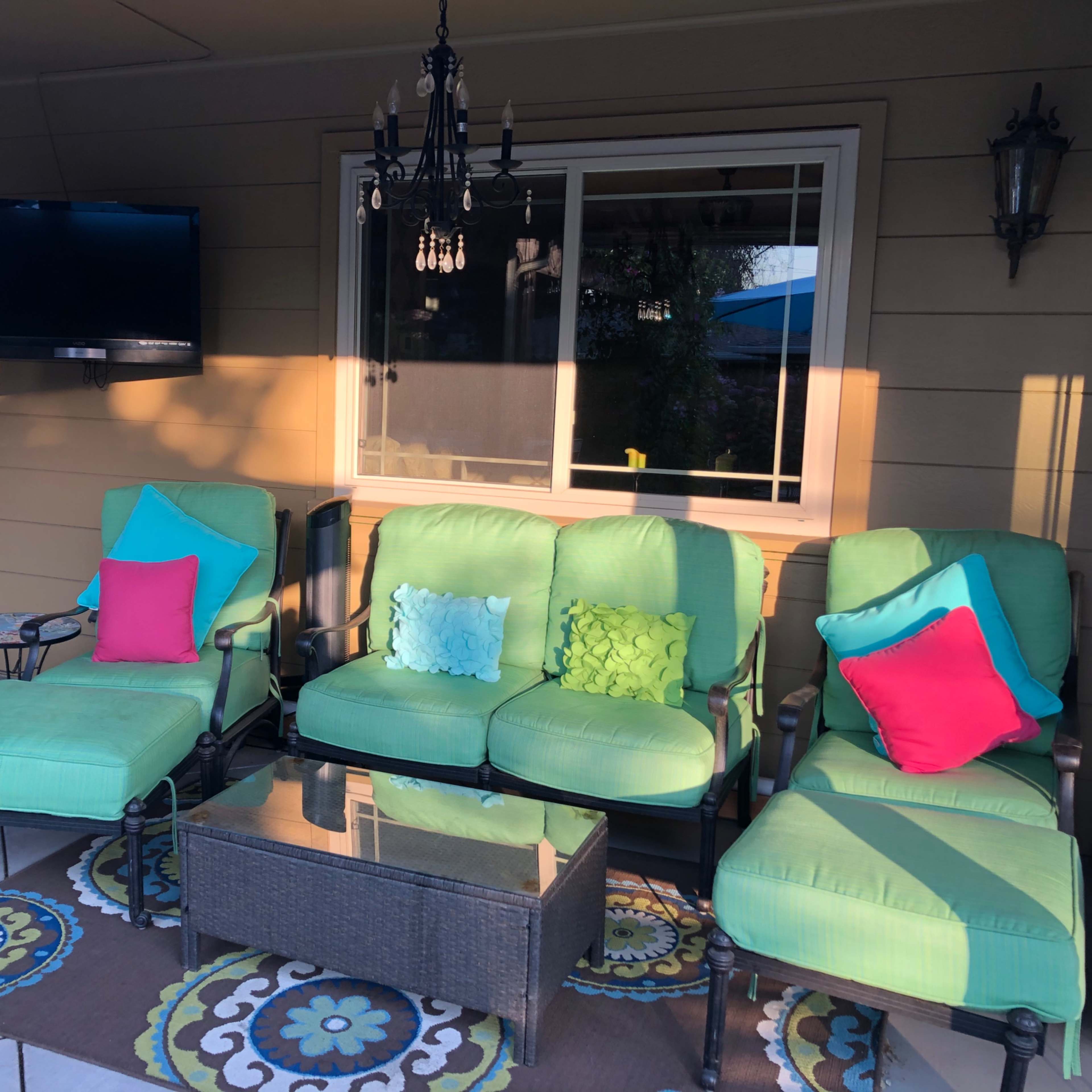 A set of green outdoor furniture with colorful pillows is arranged around a glass coffee table on a patio.