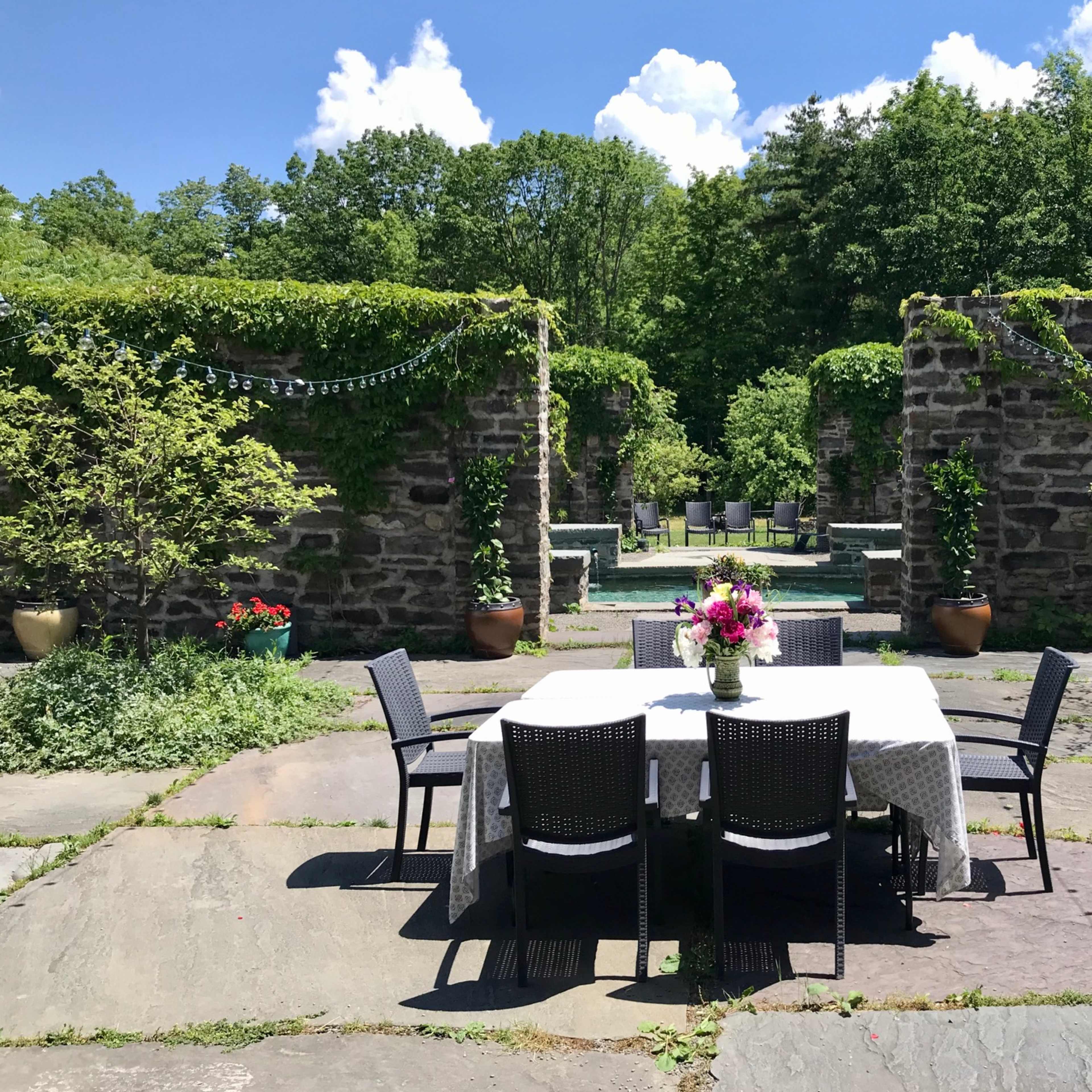 A table with a floral centerpiece is set outdoors in a stone courtyard surrounded by greenery and old stone walls.