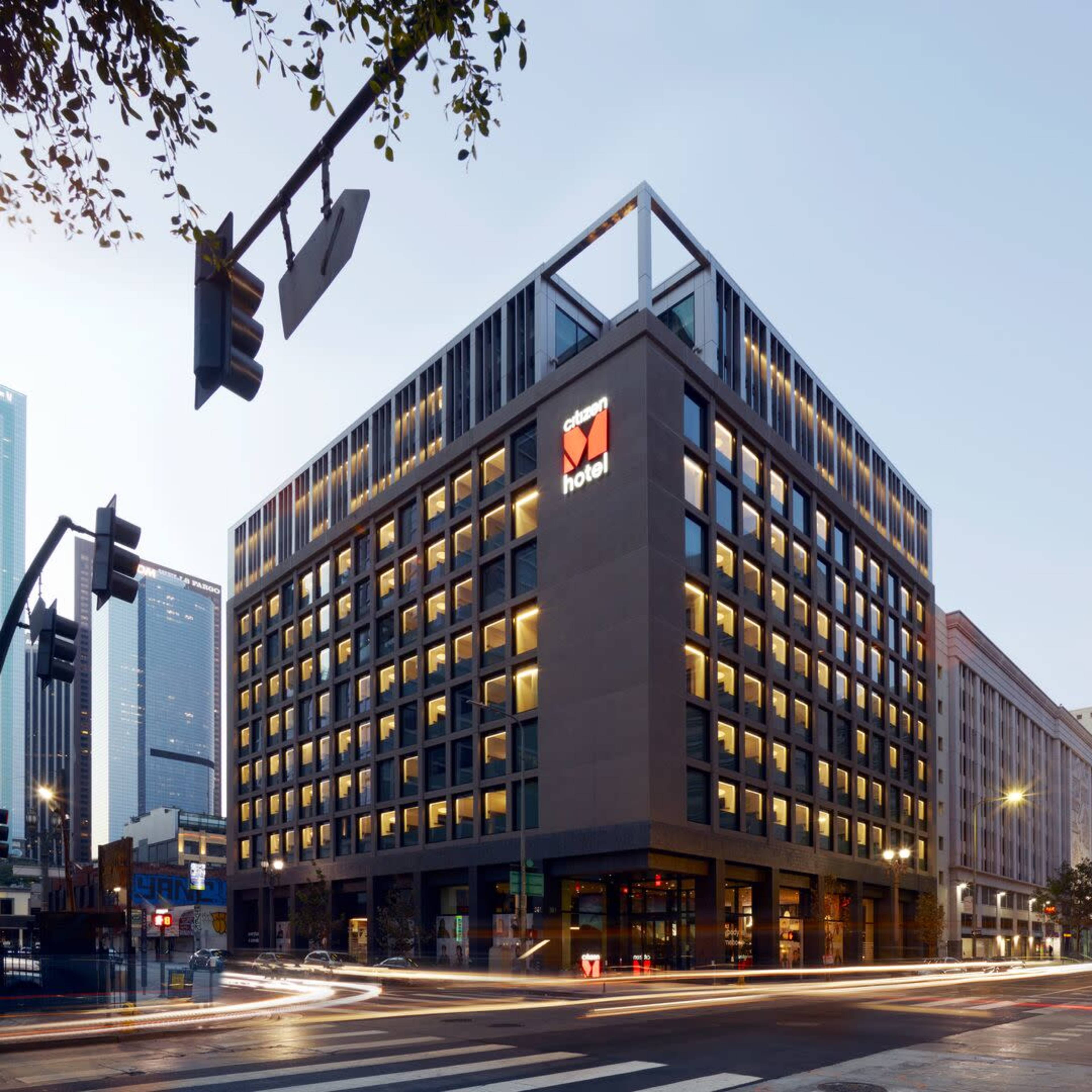 A modern hotel building at dusk, featuring a distinctive facade and illuminated windows, located at an intersection in an urban area.
