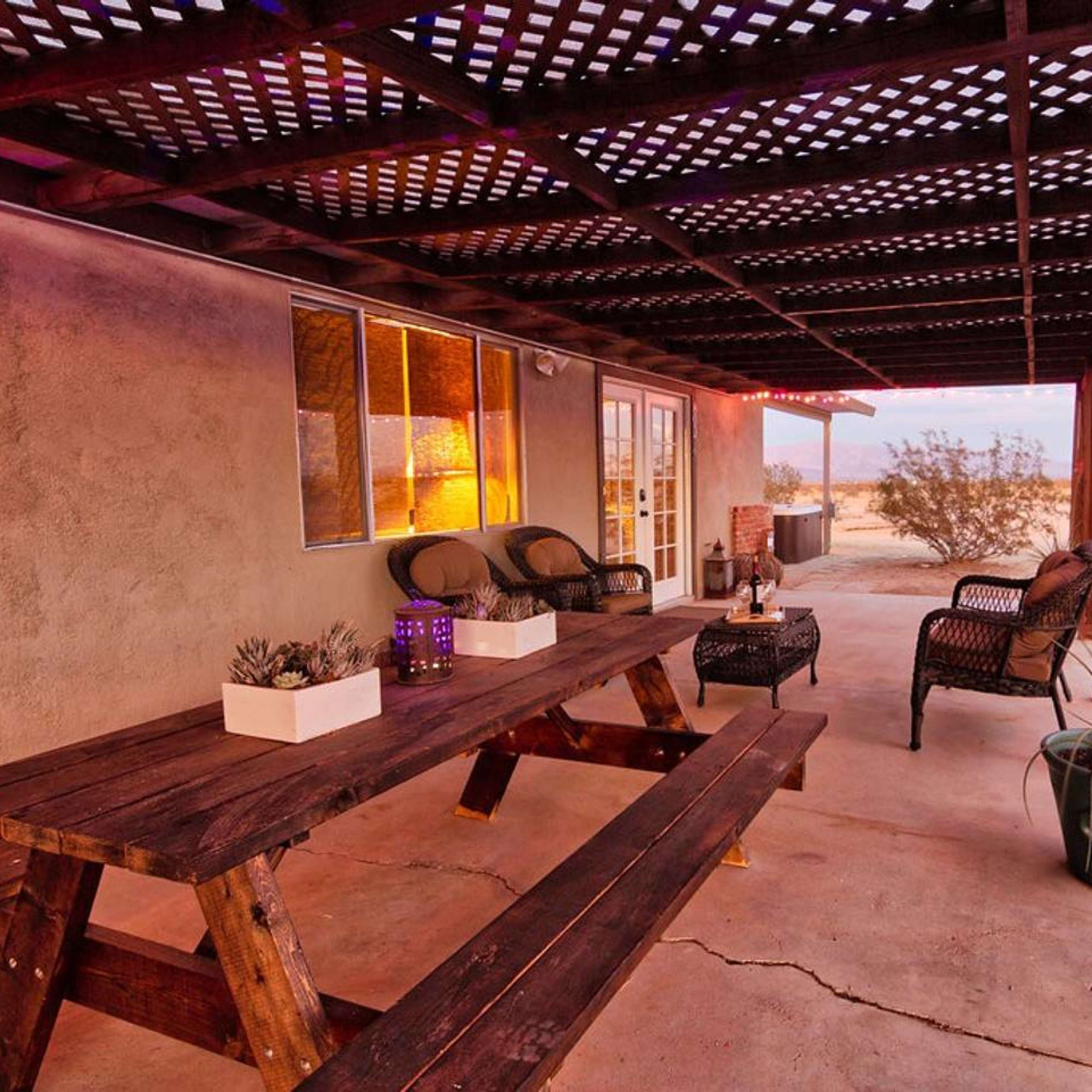 A wooden picnic table is set under a covered patio adjacent to a rustic building, with potted plants and comfortable seating nearby.