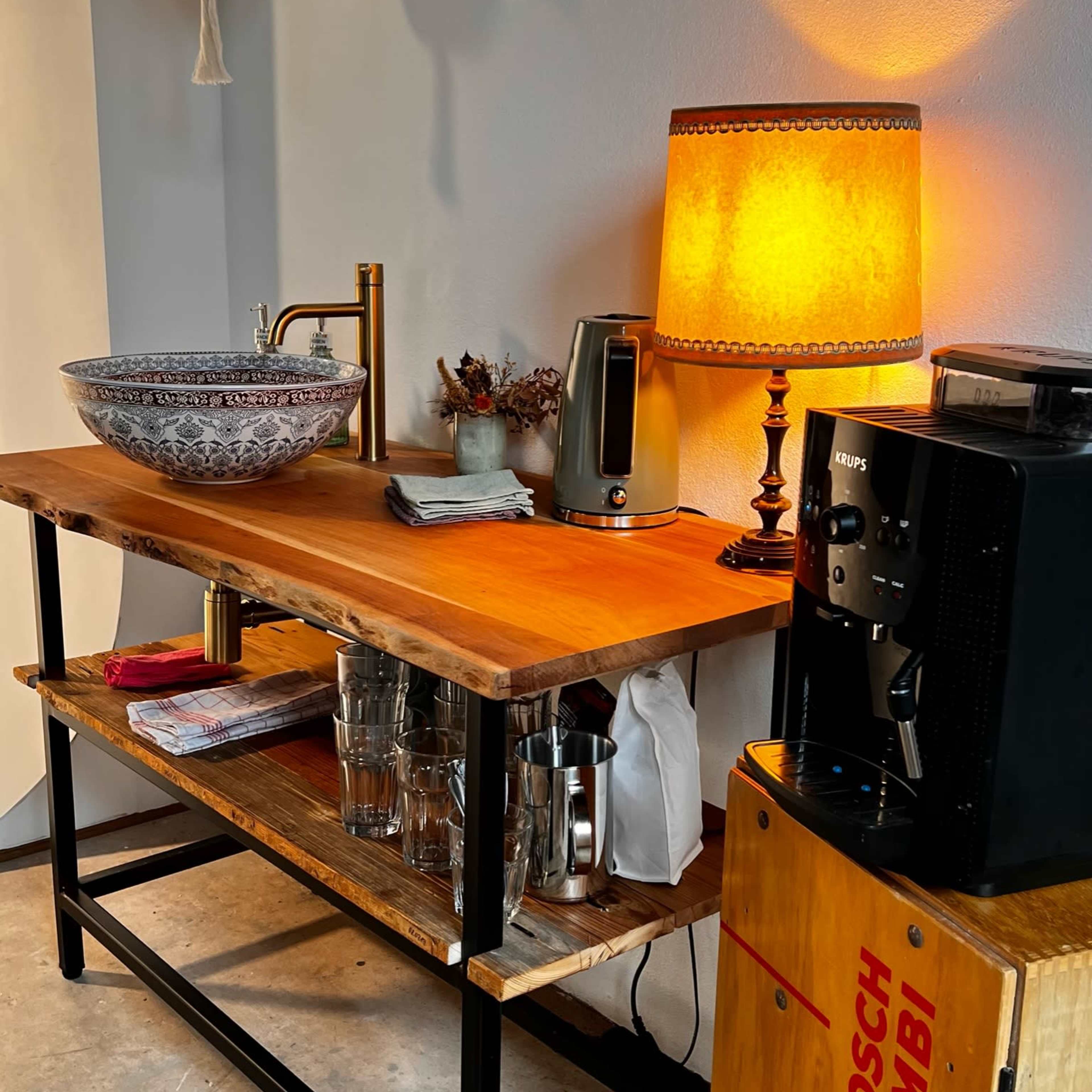 A wooden table with a decorative bowl, a kettle, a lamp, a coffee machine, and glasses, against a wall with a hanging planter.