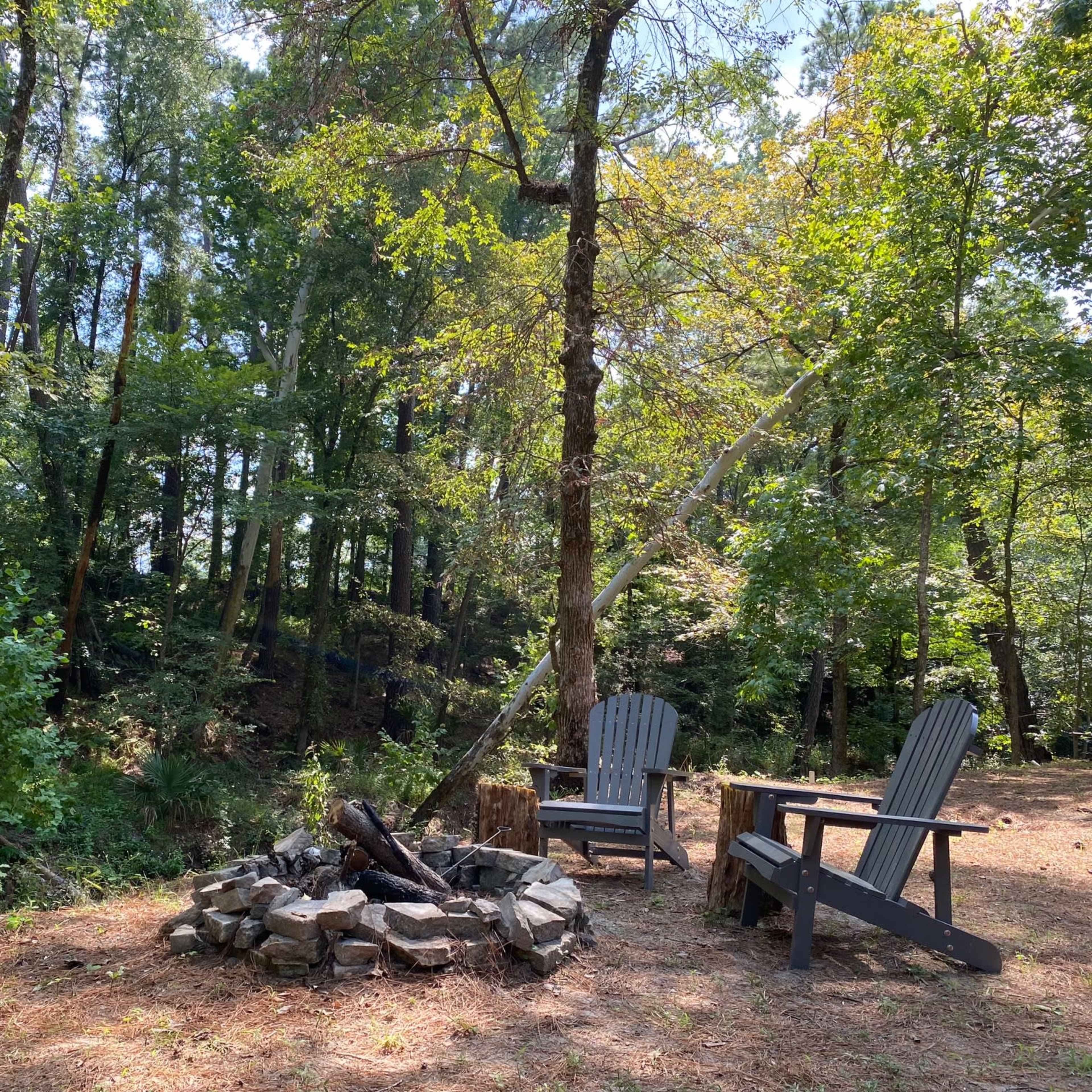 A fire pit made of stones sits in a clearing with two wooden chairs beside it, surrounded by dense trees and greenery.