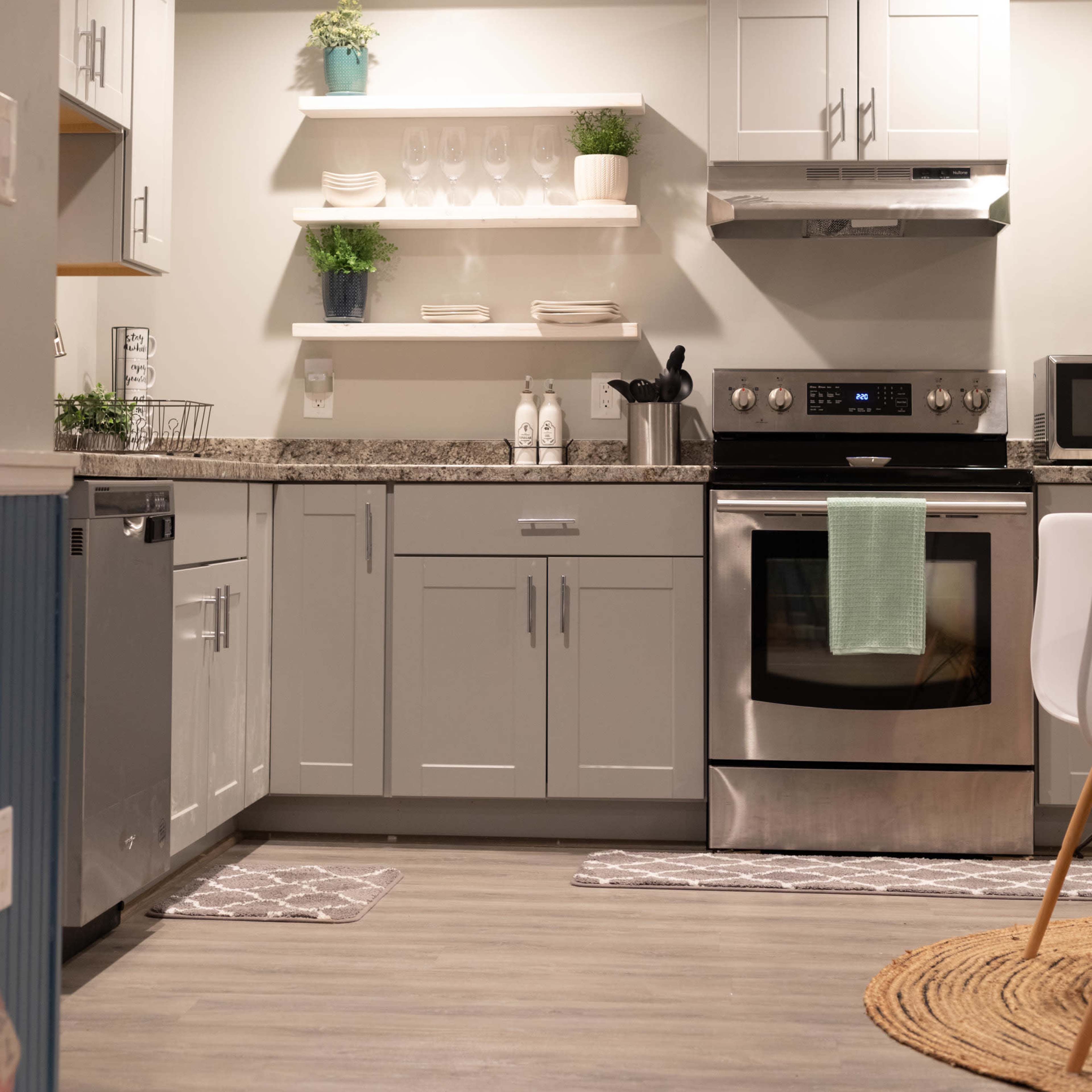 A modern kitchen with grey cabinets, a stainless steel stove, open shelving with plants, and a light-colored countertop.