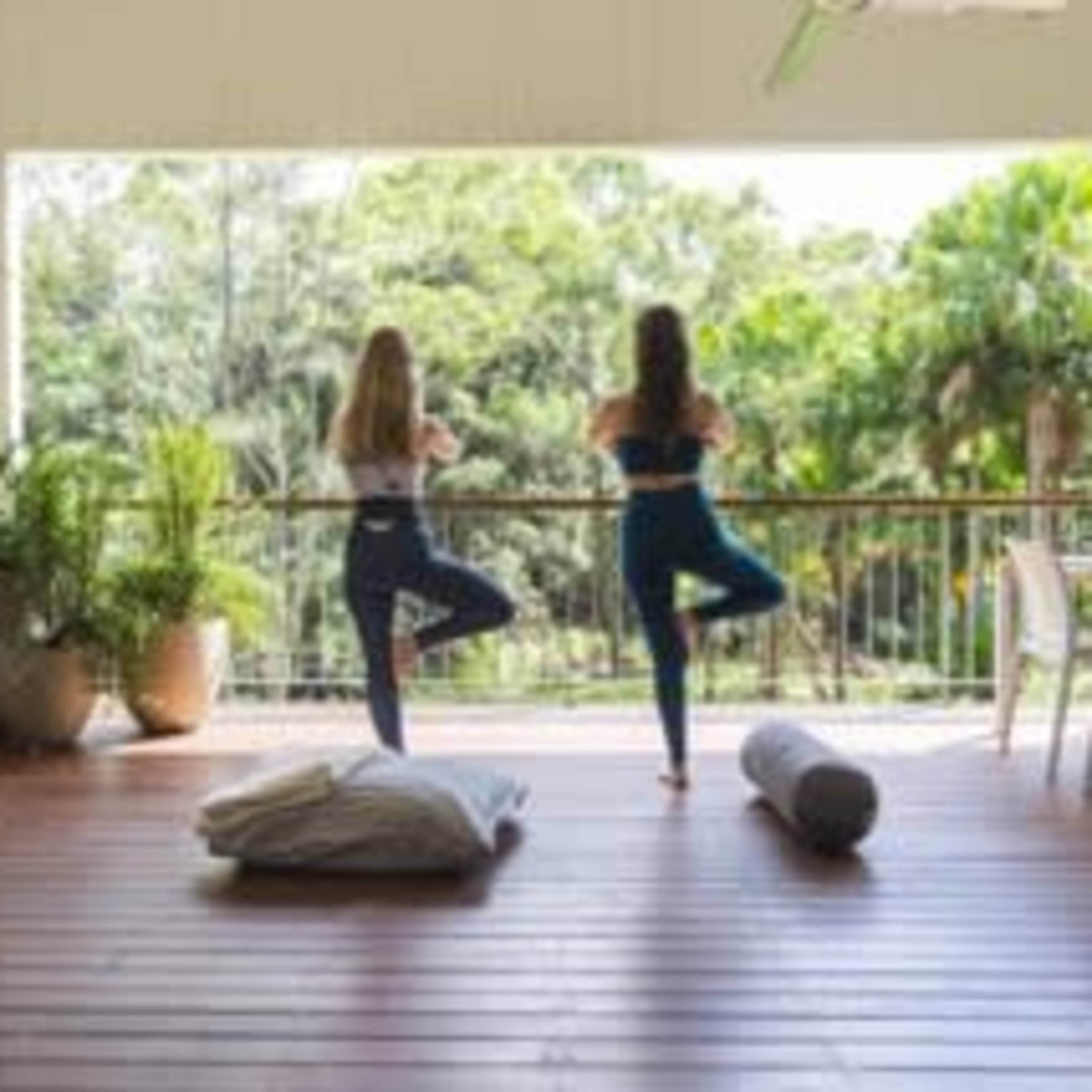 Two women practice yoga in a spacious, open area with a wooden deck overlooking a lush green landscape.
