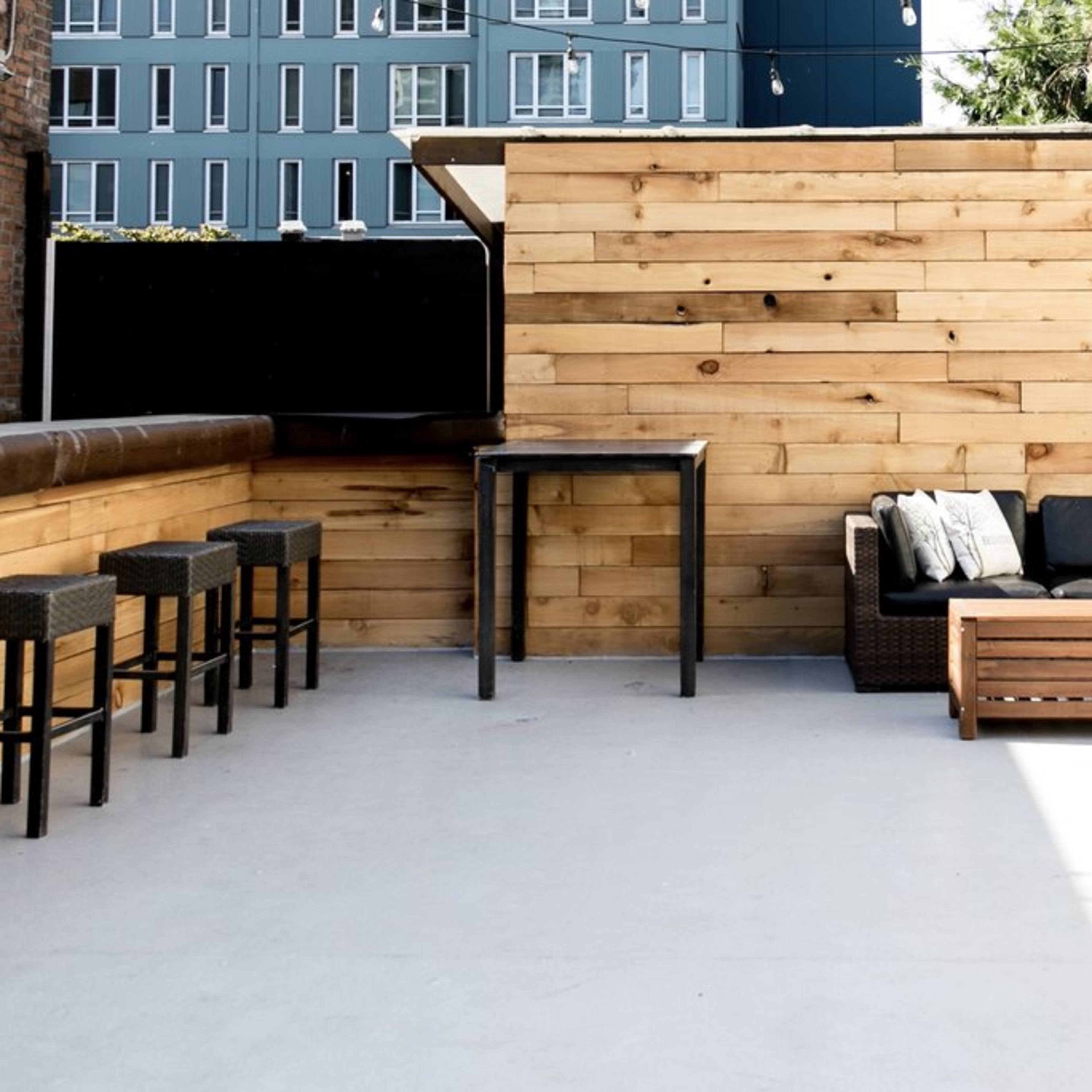 A rooftop patio featuring wooden walls, a black couch, a coffee table, and several bar stools along one side.
