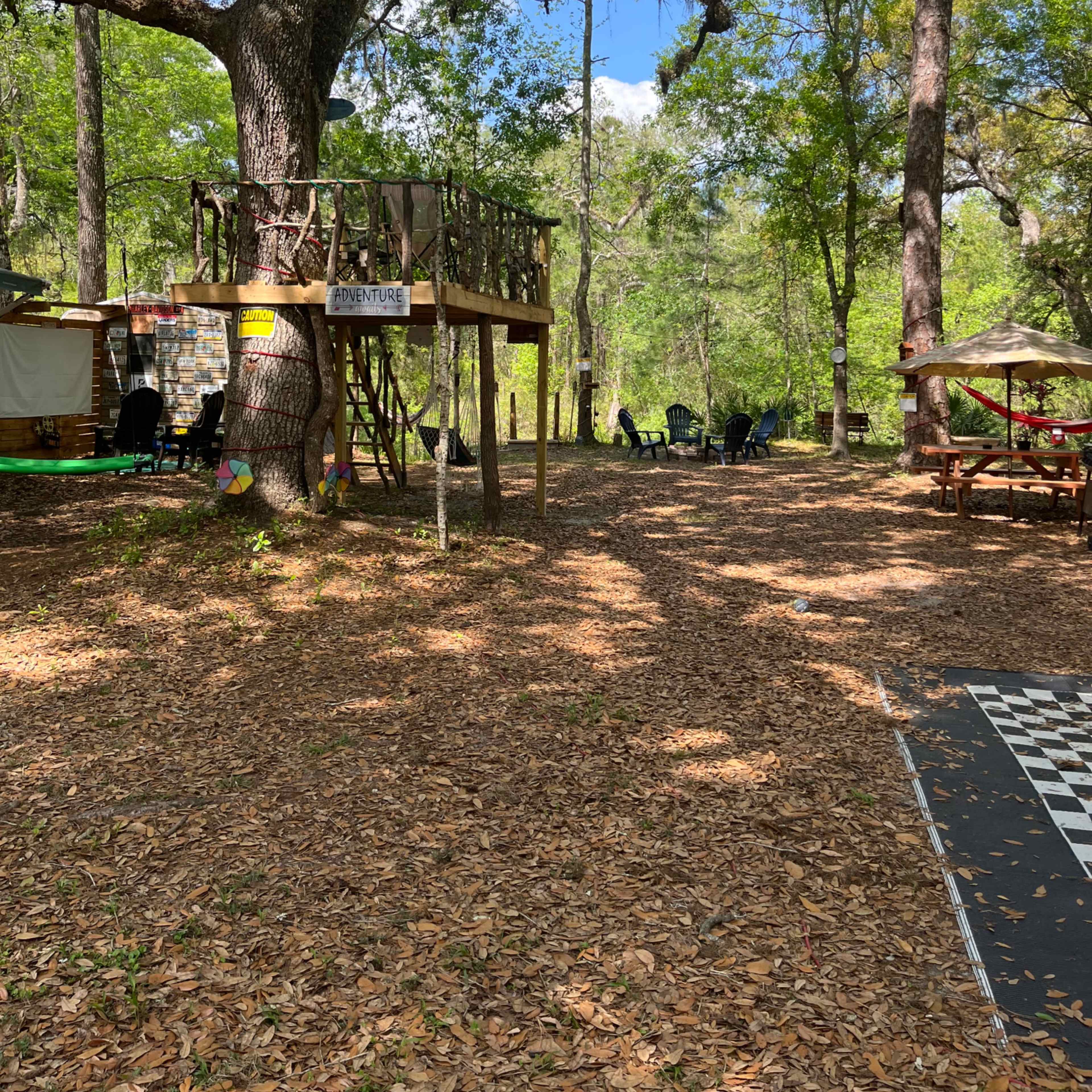 A wooded area with a treehouse on stilts, surrounded by chairs and picnic tables on a leaf-covered ground.