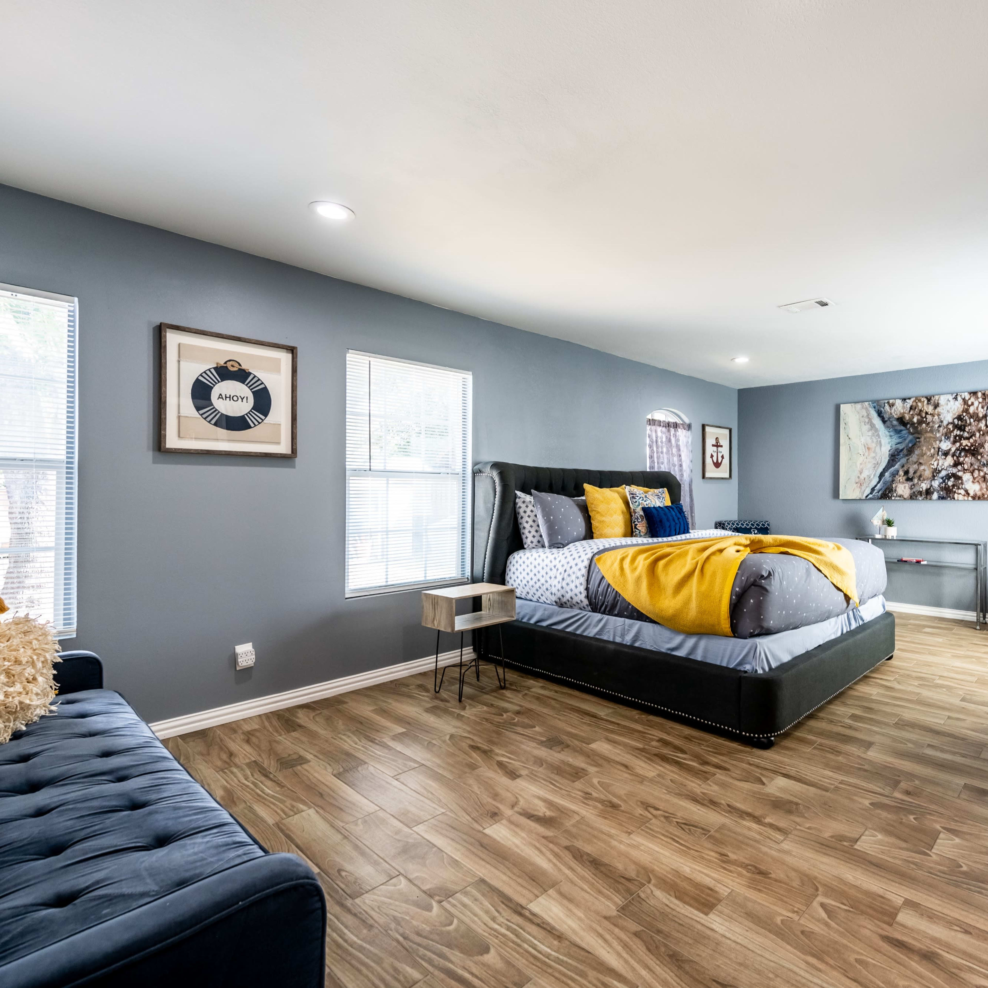 The room features a bed with yellow and gray bedding, a blue sofa, and a television mounted on the wall, with windows allowing natural light to enter.