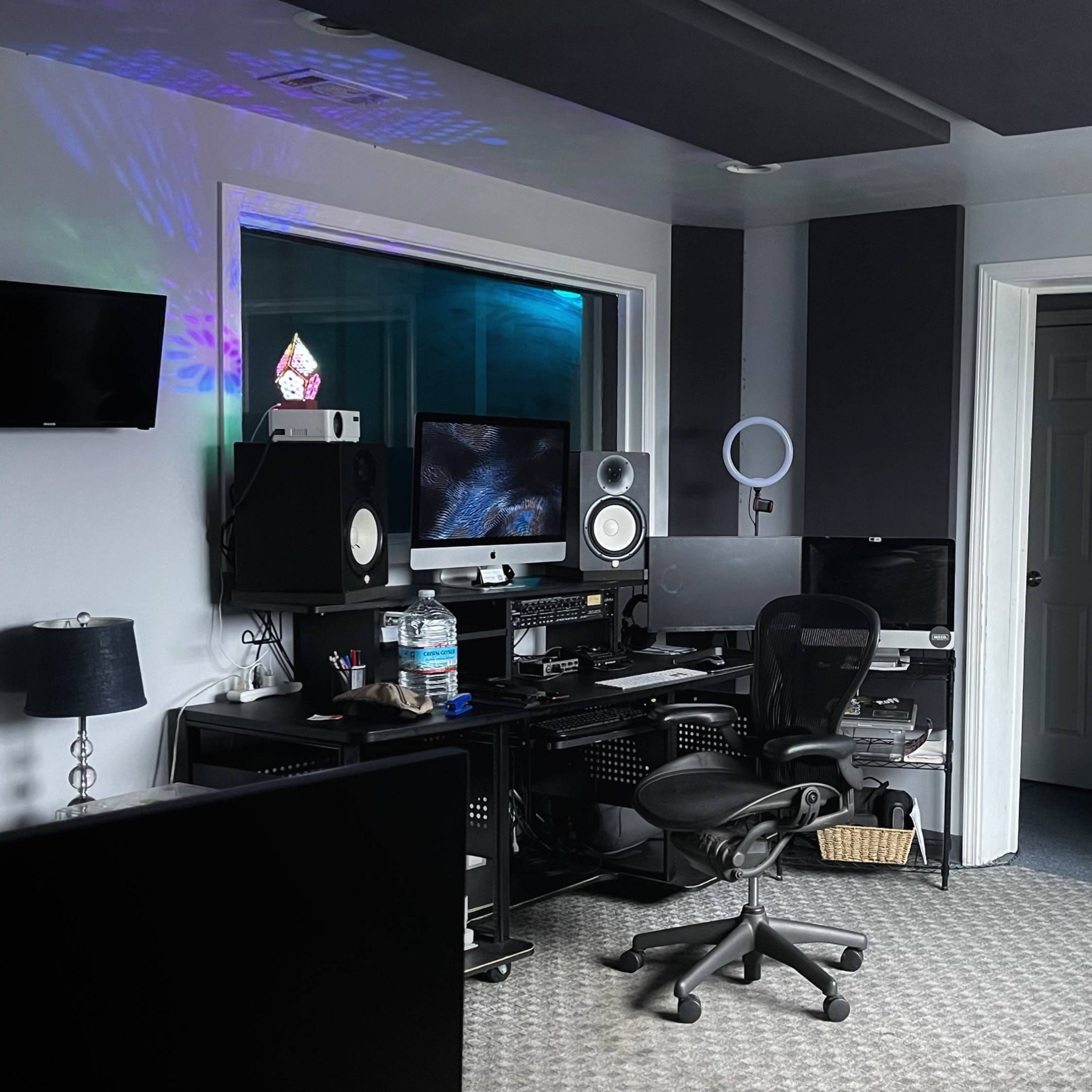 A music production studio featuring a desk with multiple computers and audio equipment, a large window, and a black couch.