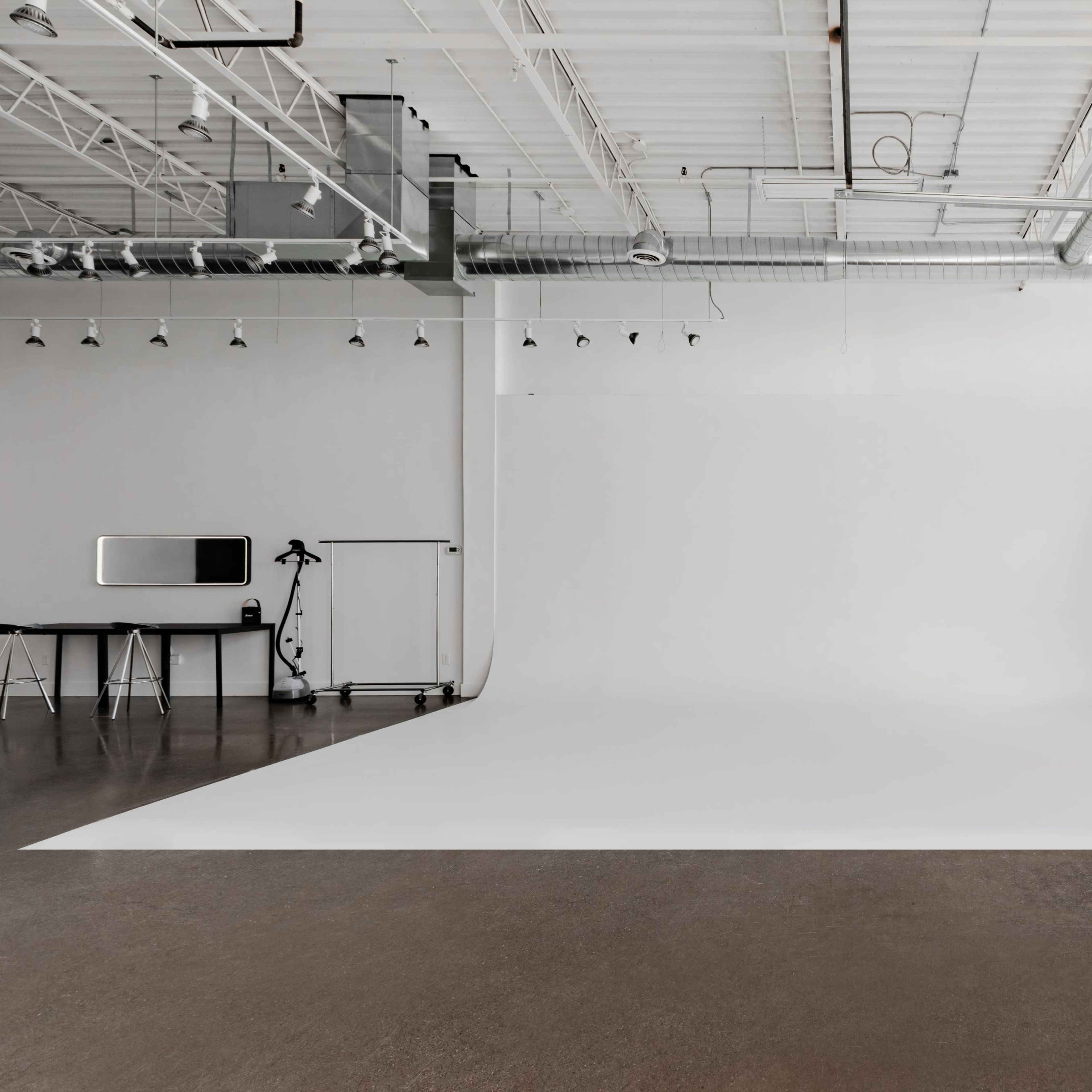 An empty photography studio with a white backdrop, a black table, and a tripod on a polished concrete floor.