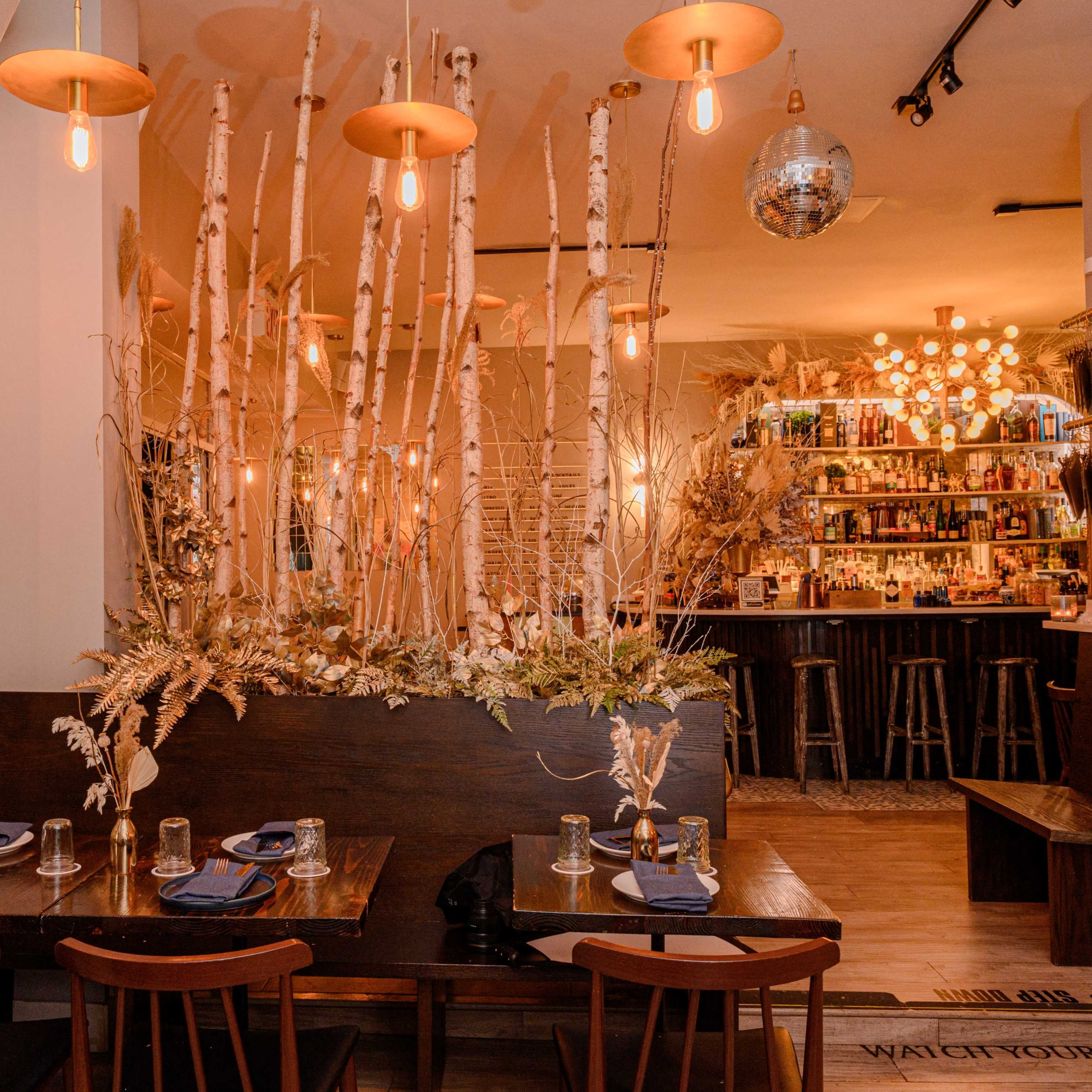 A stylish restaurant interior featuring wooden tables, hanging lights, and decorative branches, with a bar in the background.