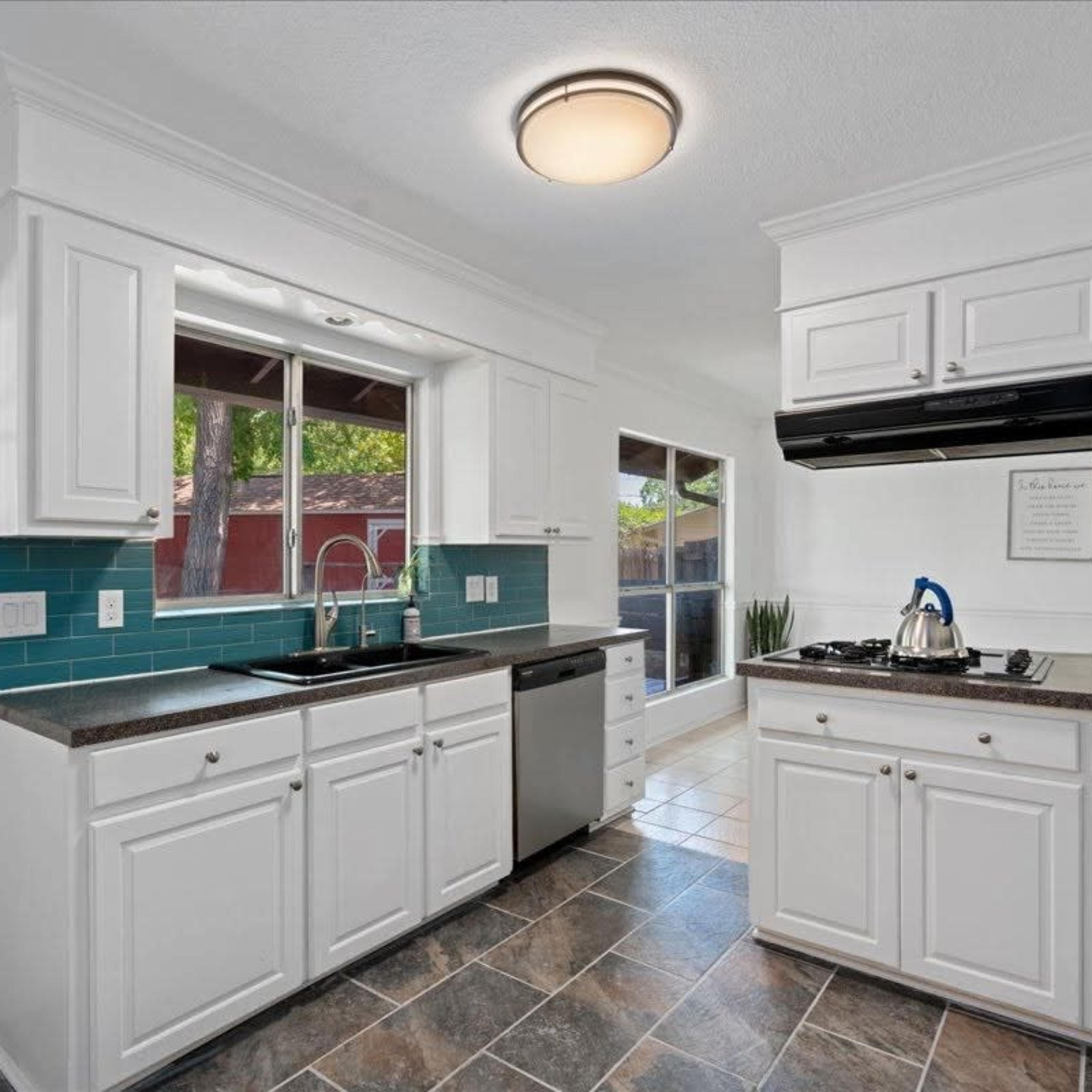 A modern kitchen with white cabinetry, a dark countertop, stainless steel appliances, and tiled flooring, featuring a window above the sink and a door leading outside.