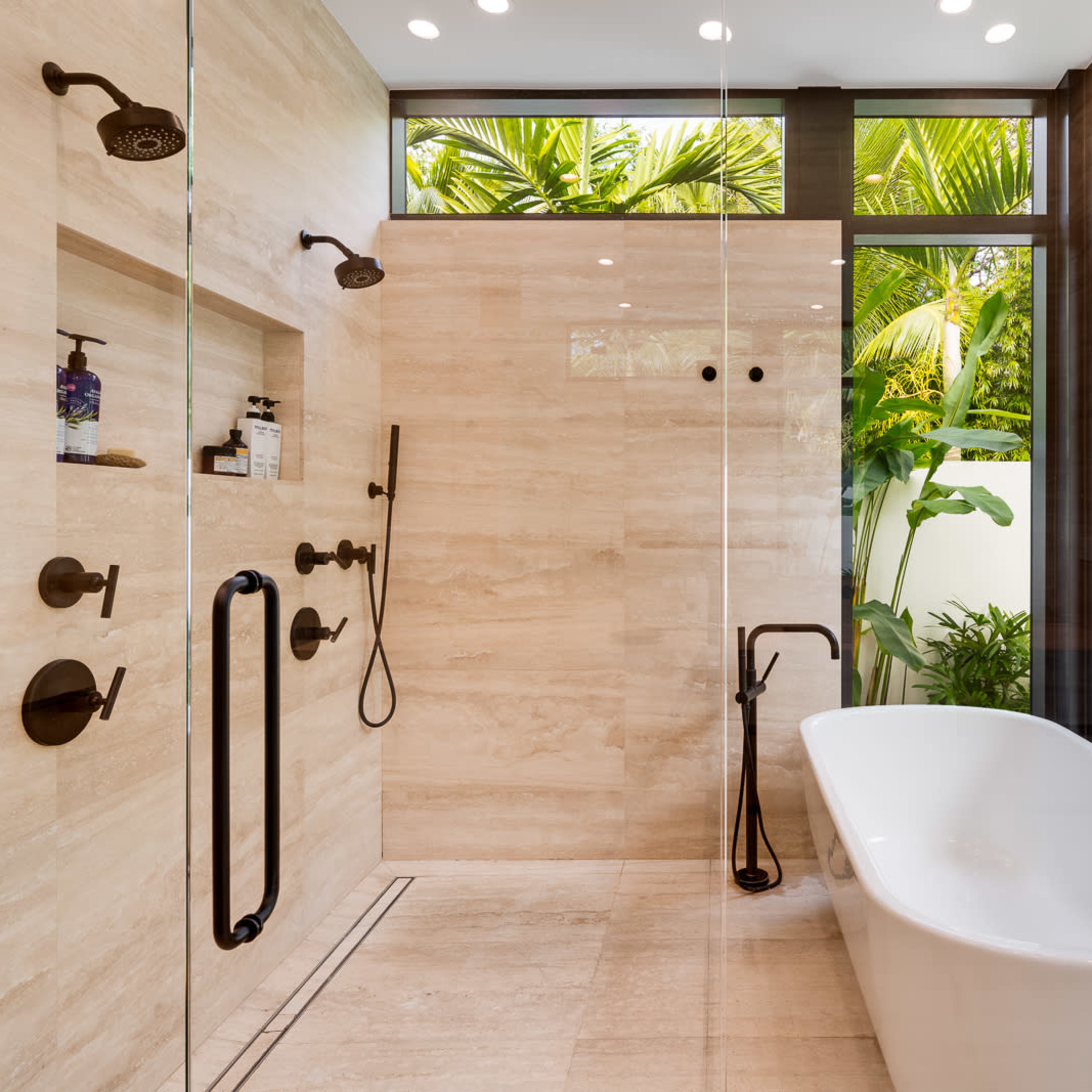 A modern bathroom featuring a glass shower with multiple showerheads, a freestanding white bathtub, and tropical plants visible through large windows.