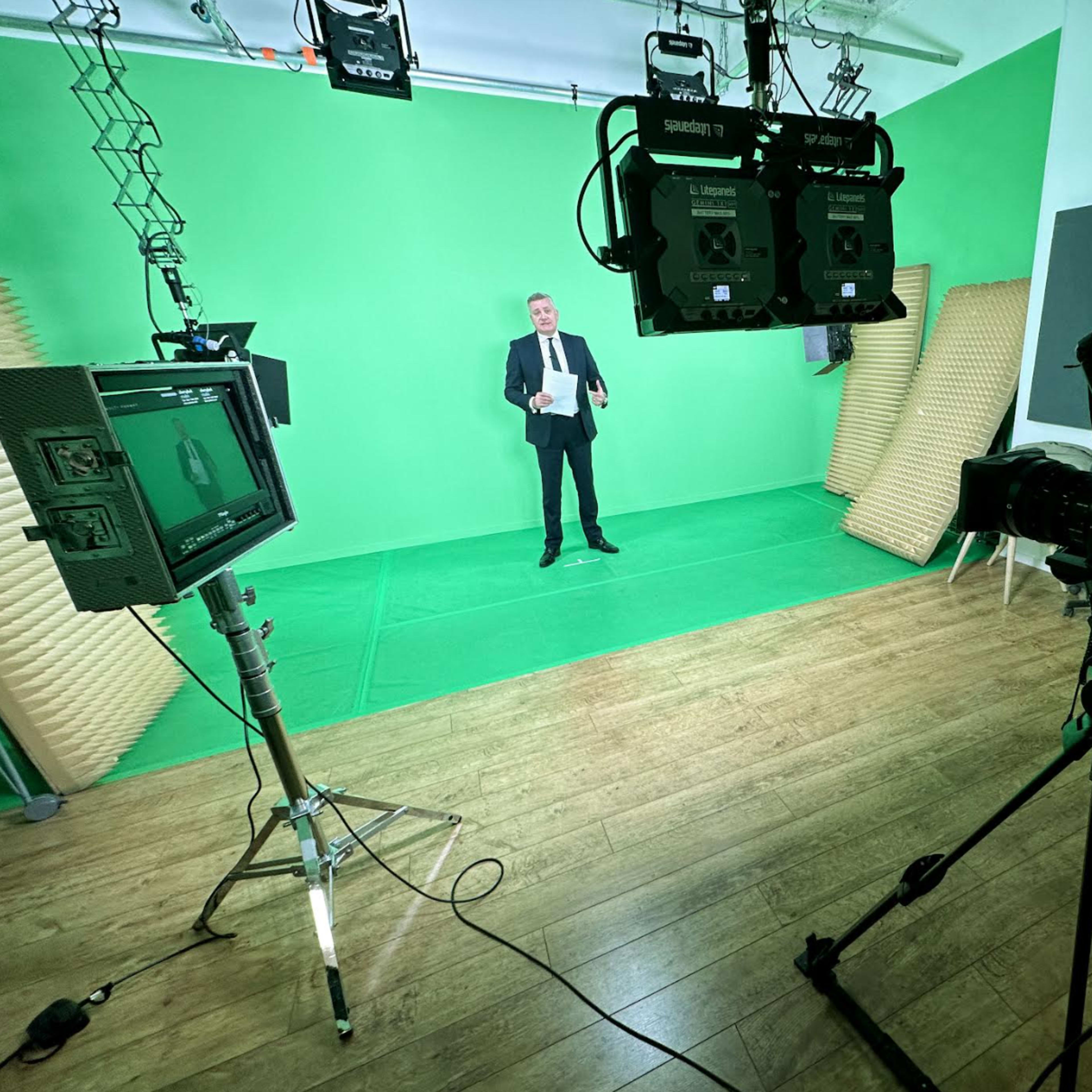 A man in a suit stands on a green studio backdrop, flanked by camera equipment and lighting fixtures.