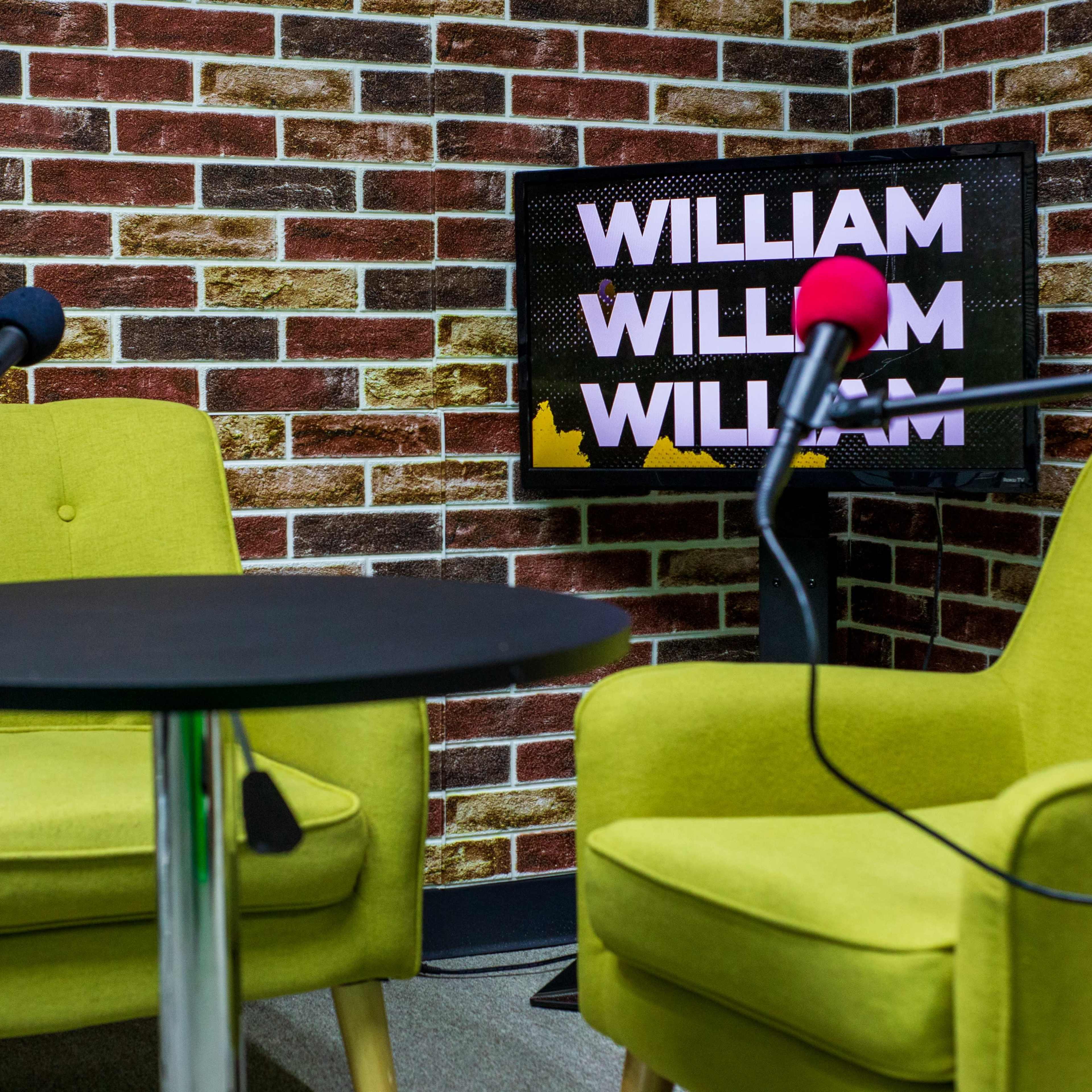 Two green chairs facing a black table with a microphone on each side, set against a brick-patterned wall and a monitor displaying the name "WILLIAM."