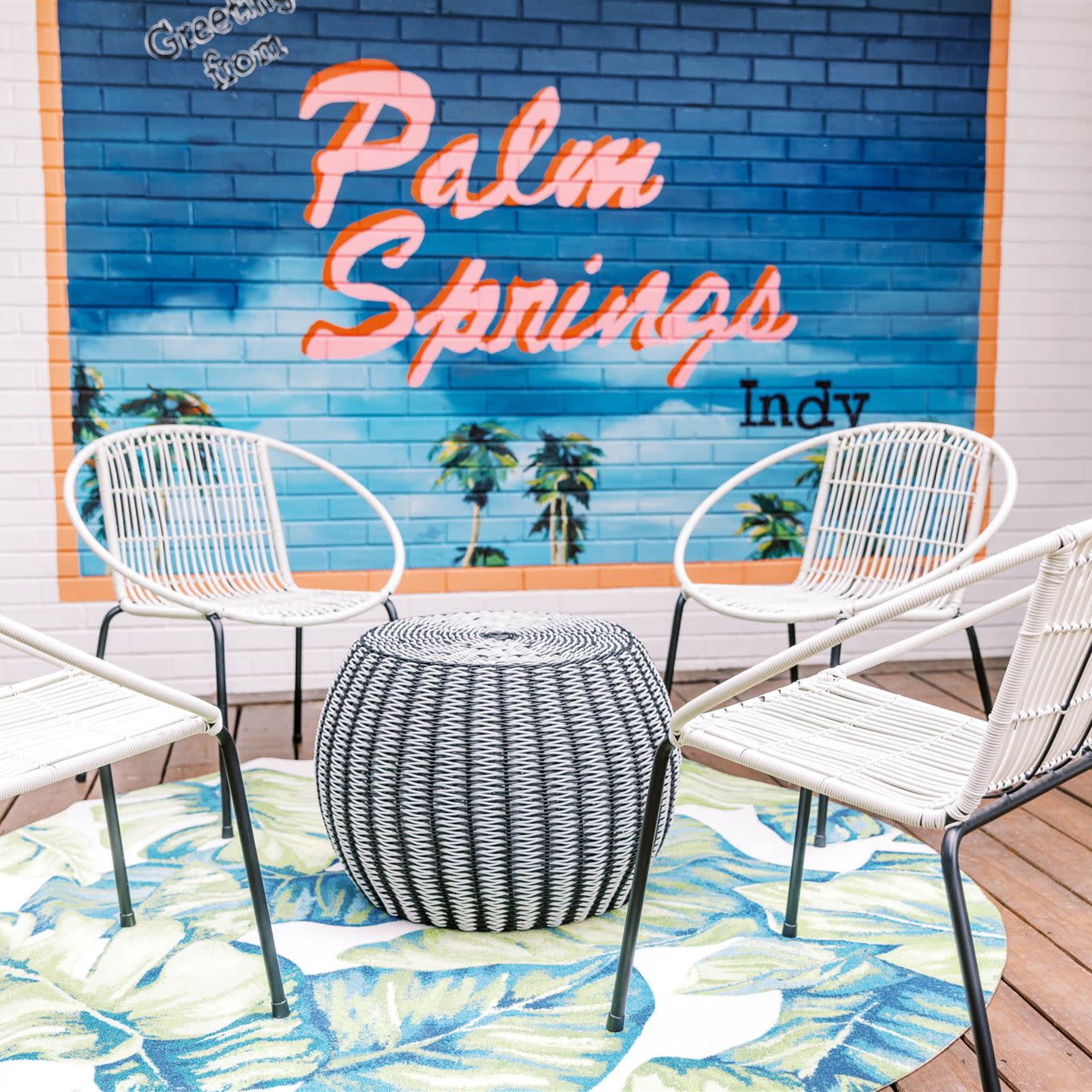 A patio area with four chairs arranged around a round table on a circular tropical-themed rug, set against a mural that reads "Palm Springs" on a blue brick wall.