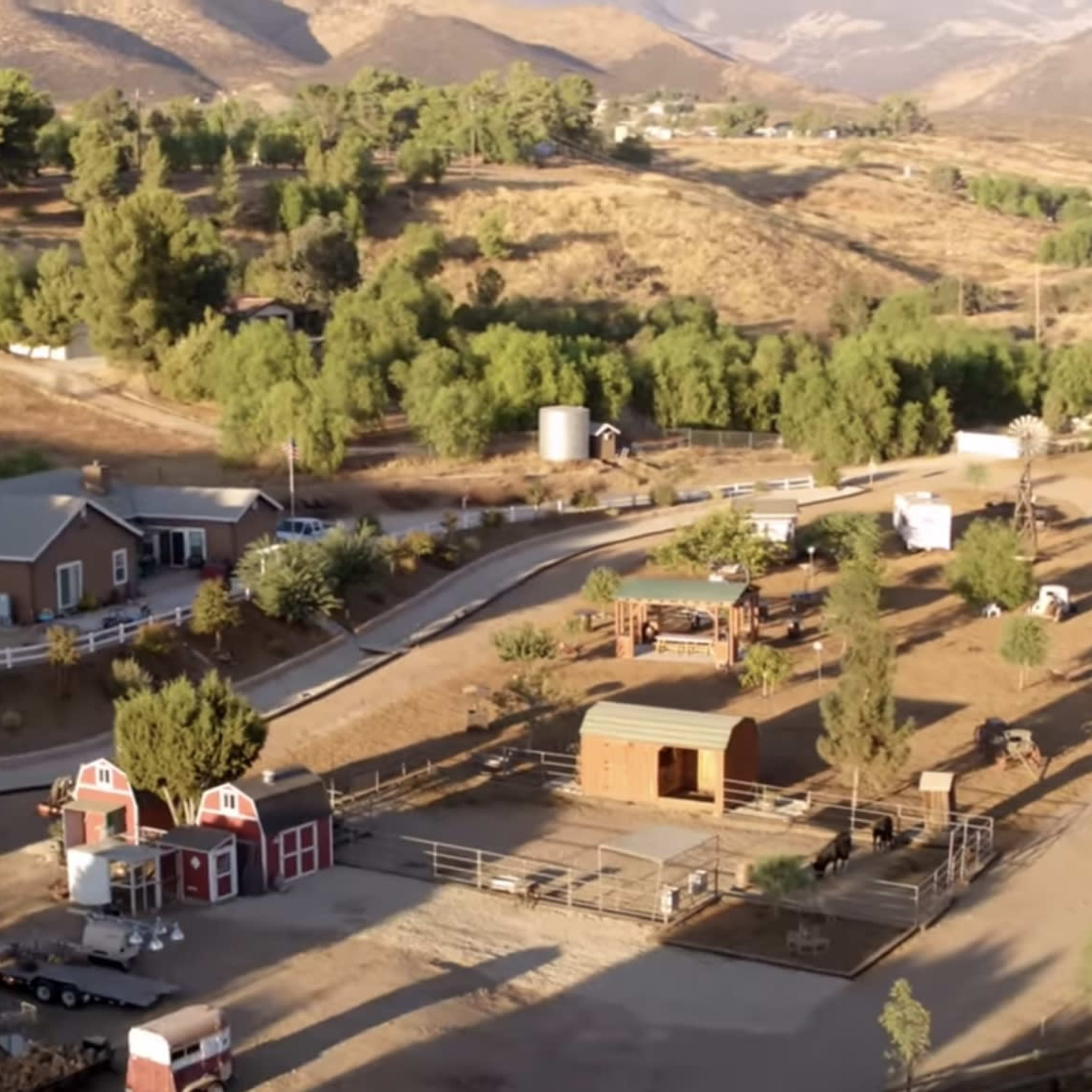 A rural landscape featuring a cluster of houses, a barn, and several paddocks surrounded by hills and trees.