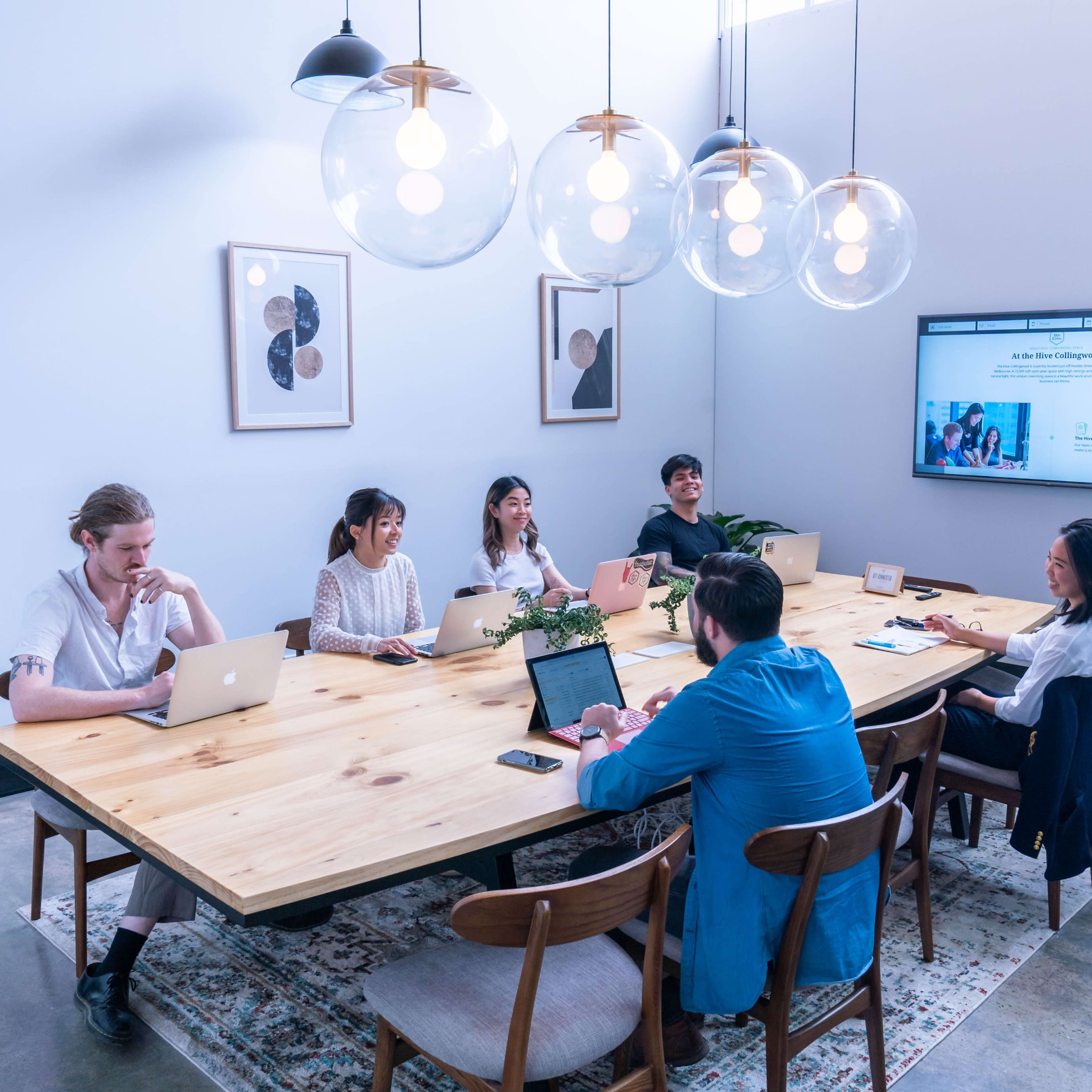 A group of six people sit around a large wooden conference table with laptops, engaged in a meeting in a well-lit room.