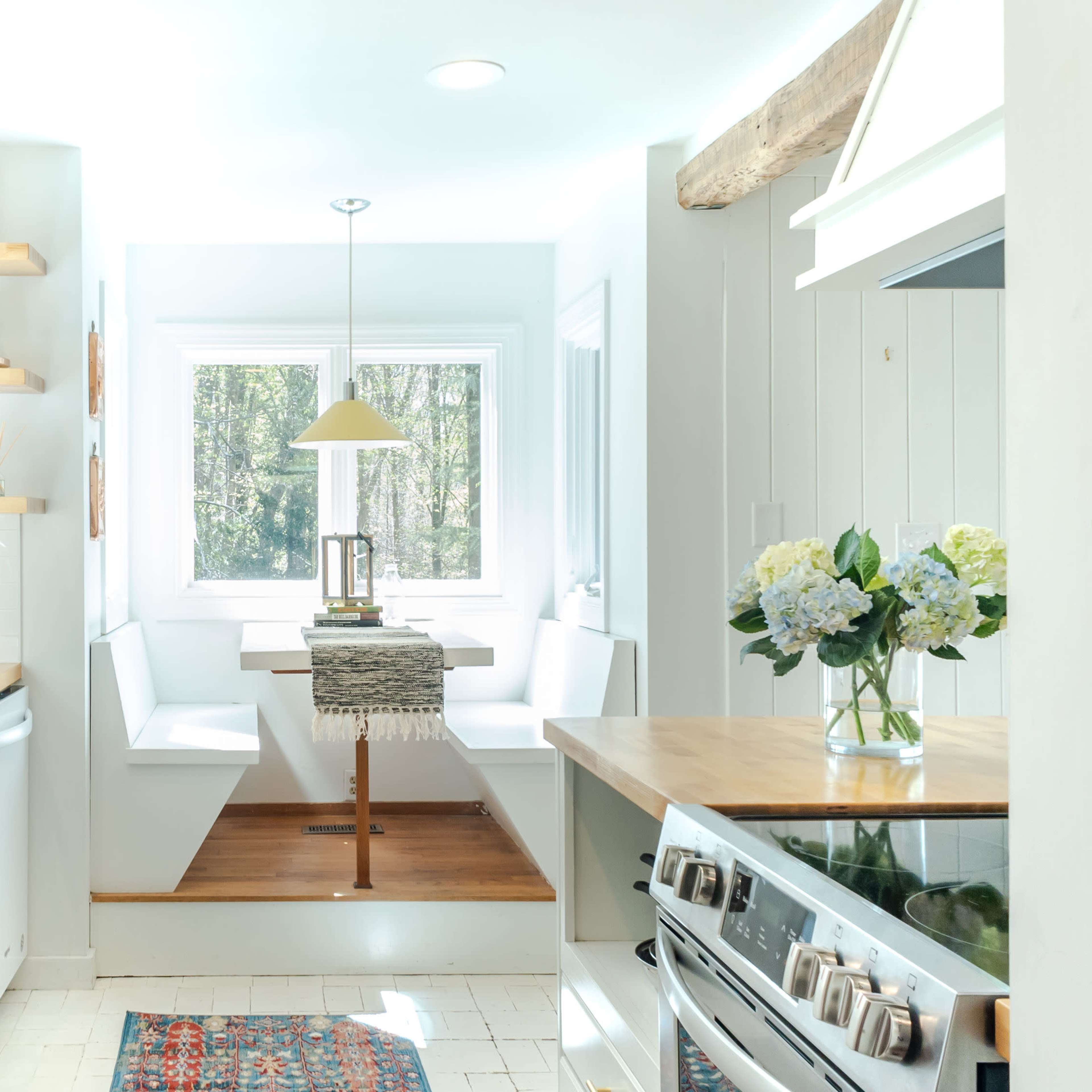 A modern kitchen features white cabinetry, a central wooden dining table with built-in seating, and large windows allowing natural light to enter.