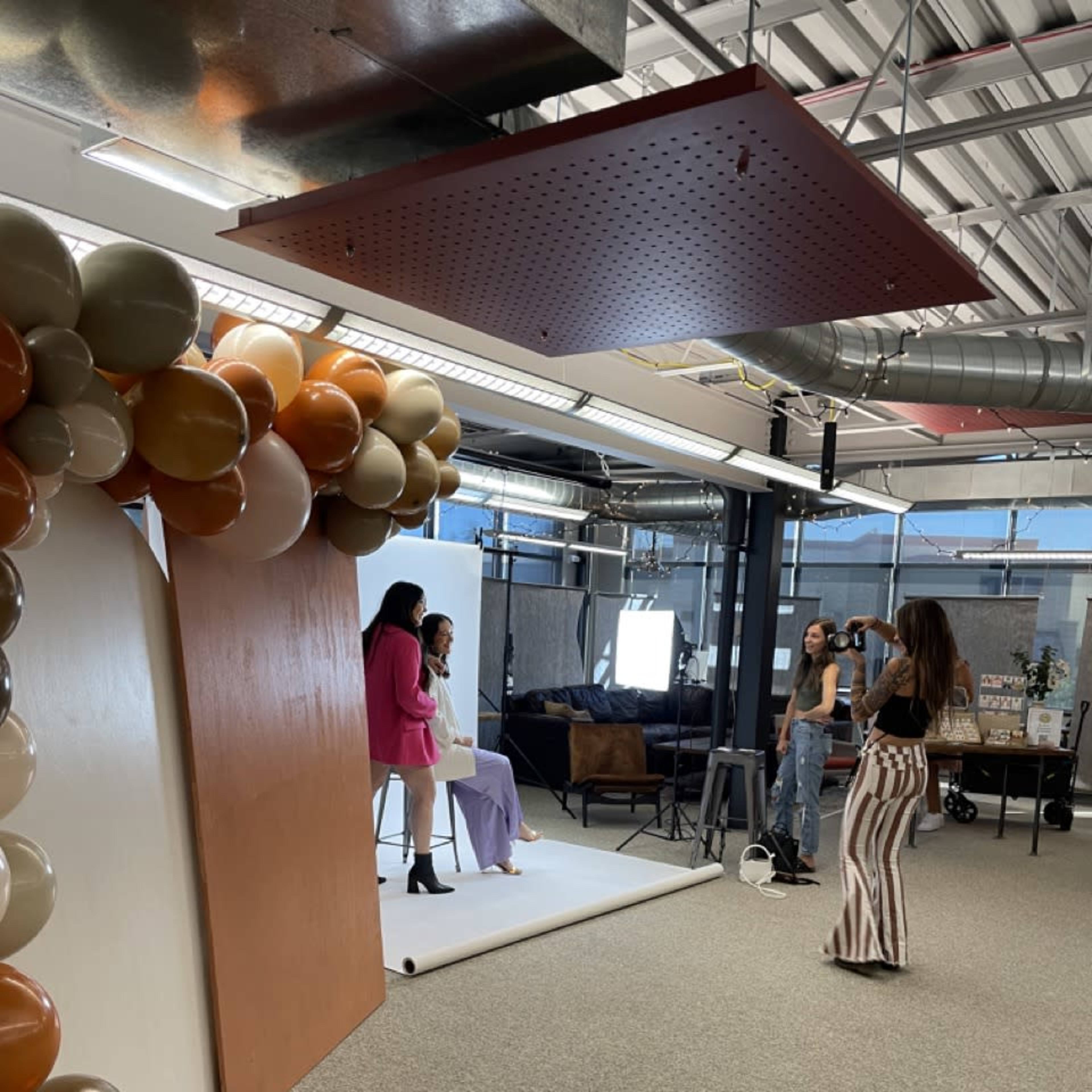 Two women pose for a photo shoot in an office space decorated with balloons and a backdrop, while a photographer captures the scene.
