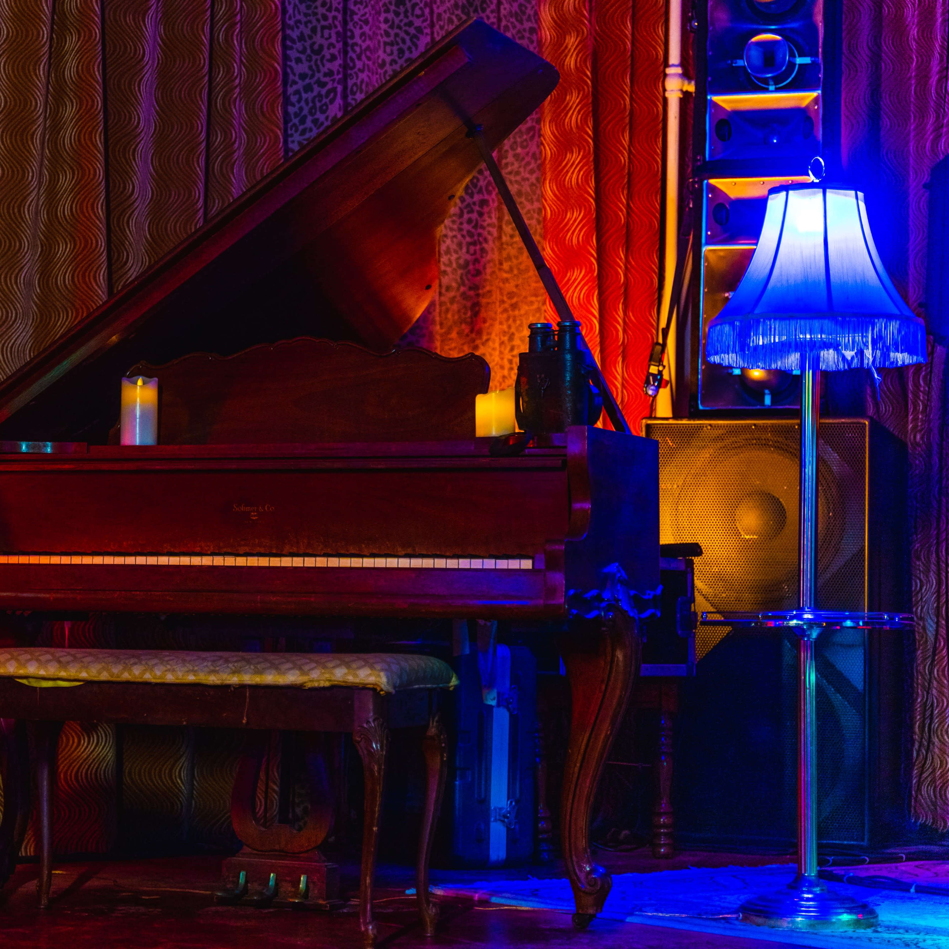 A grand piano with its lid open is positioned next to a plant, a lamp, and speakers against a backdrop of patterned drapes.