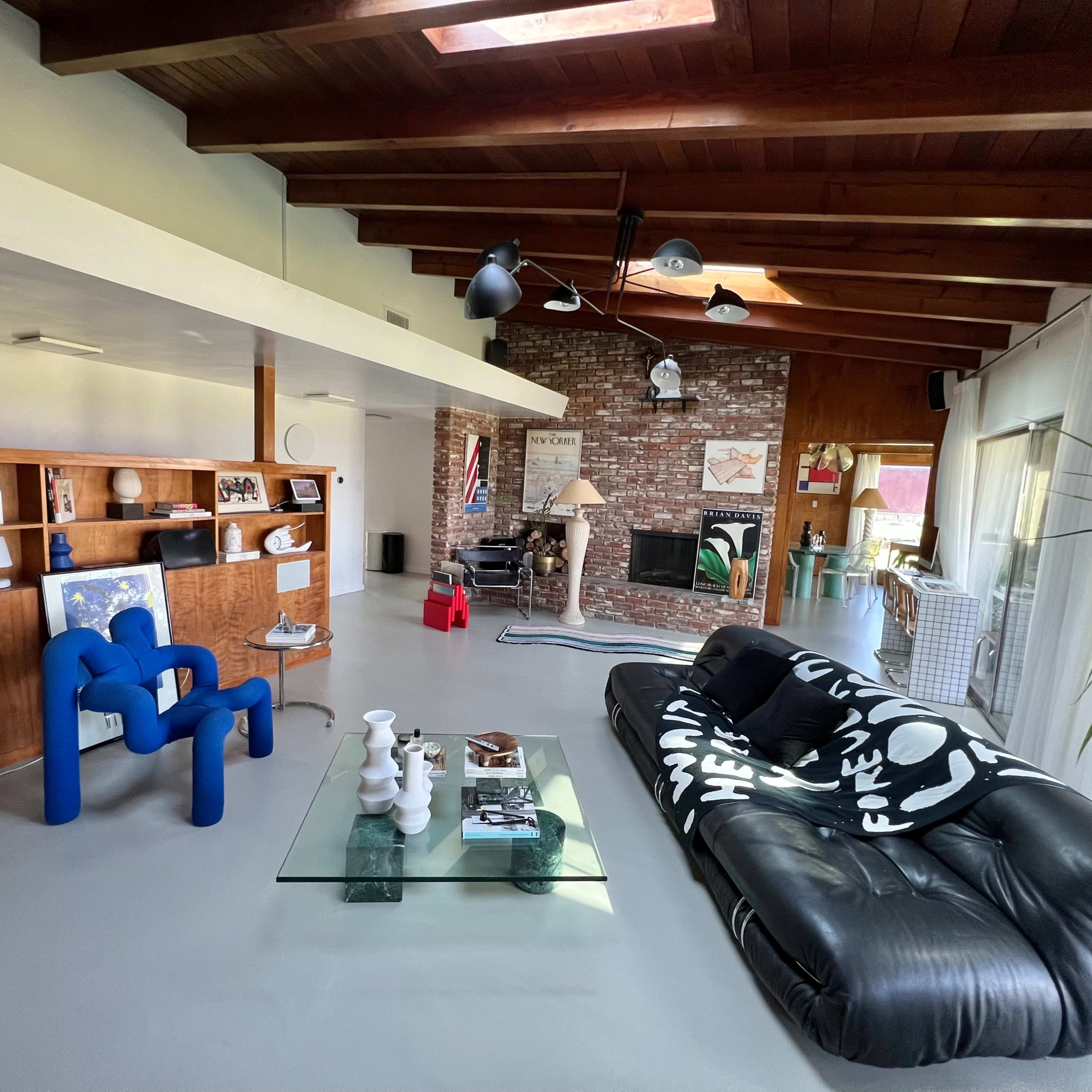 A spacious living room with a mix of modern furniture, including a blue chair, a black sofa with white patterns, and a glass coffee table, surrounded by wooden beams and a brick wall.