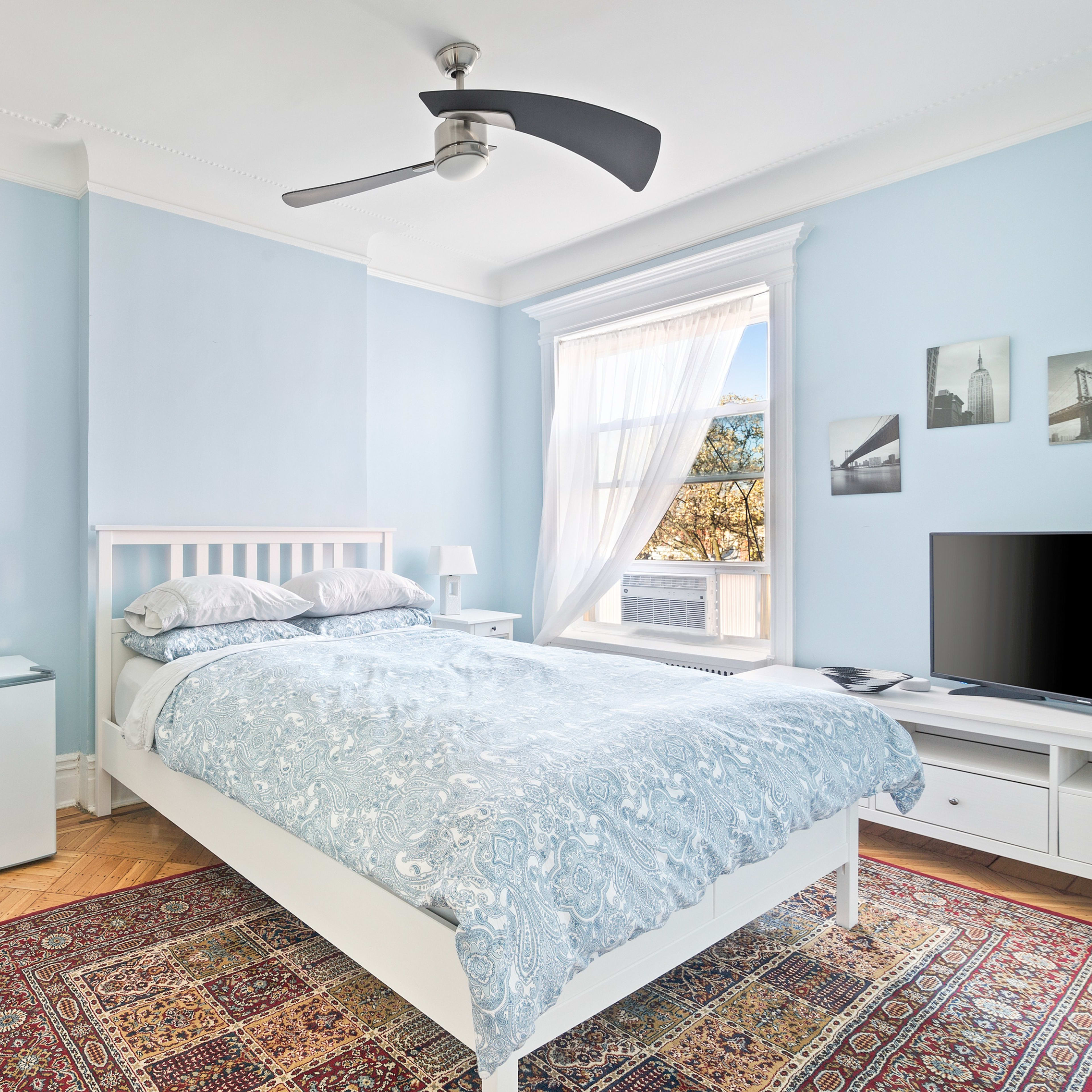 A light blue bedroom features a white bed with a patterned comforter, a nightstand, a television, and a large window with sheer curtains.