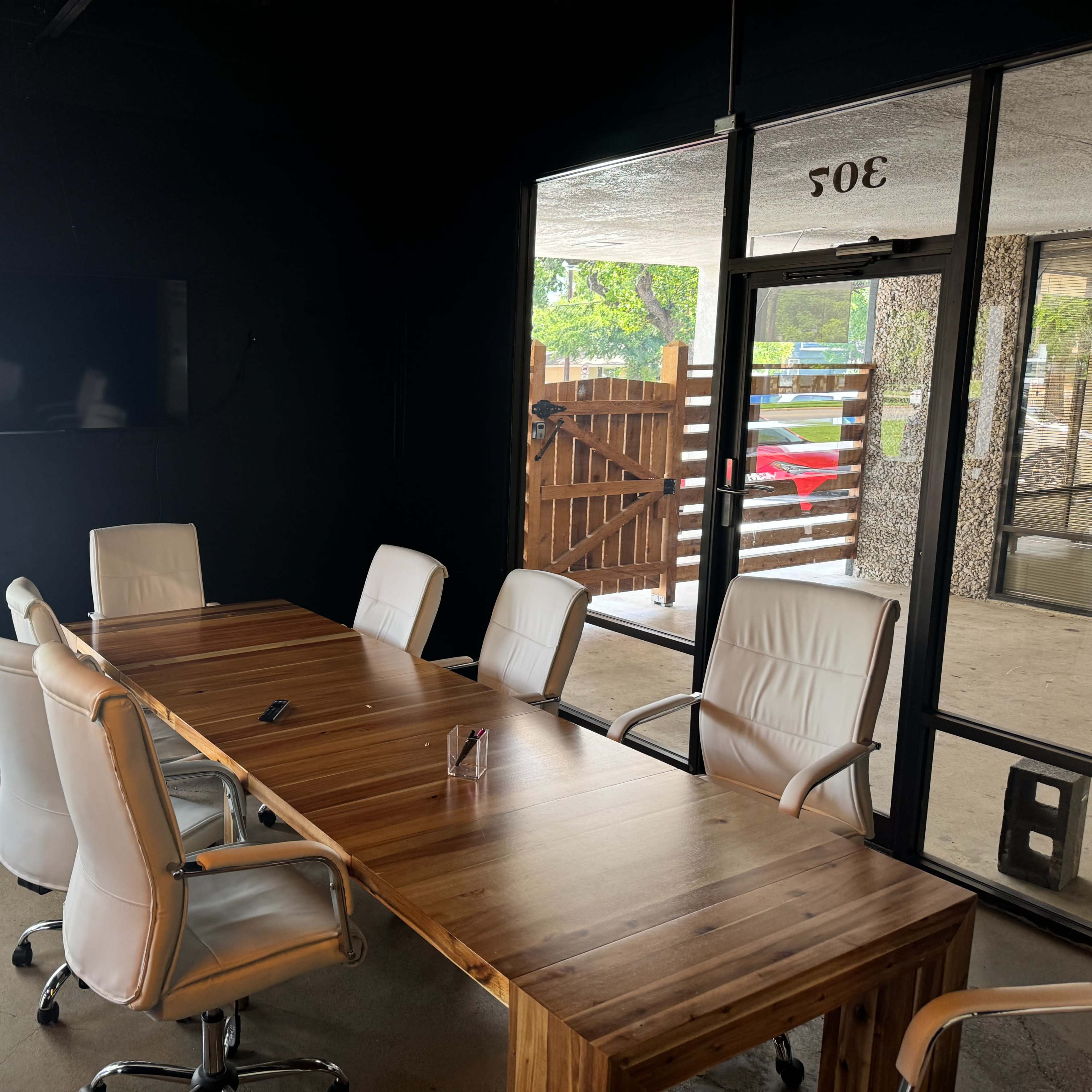 A conference room features a wooden table surrounded by white chairs, with large glass windows letting in natural light.