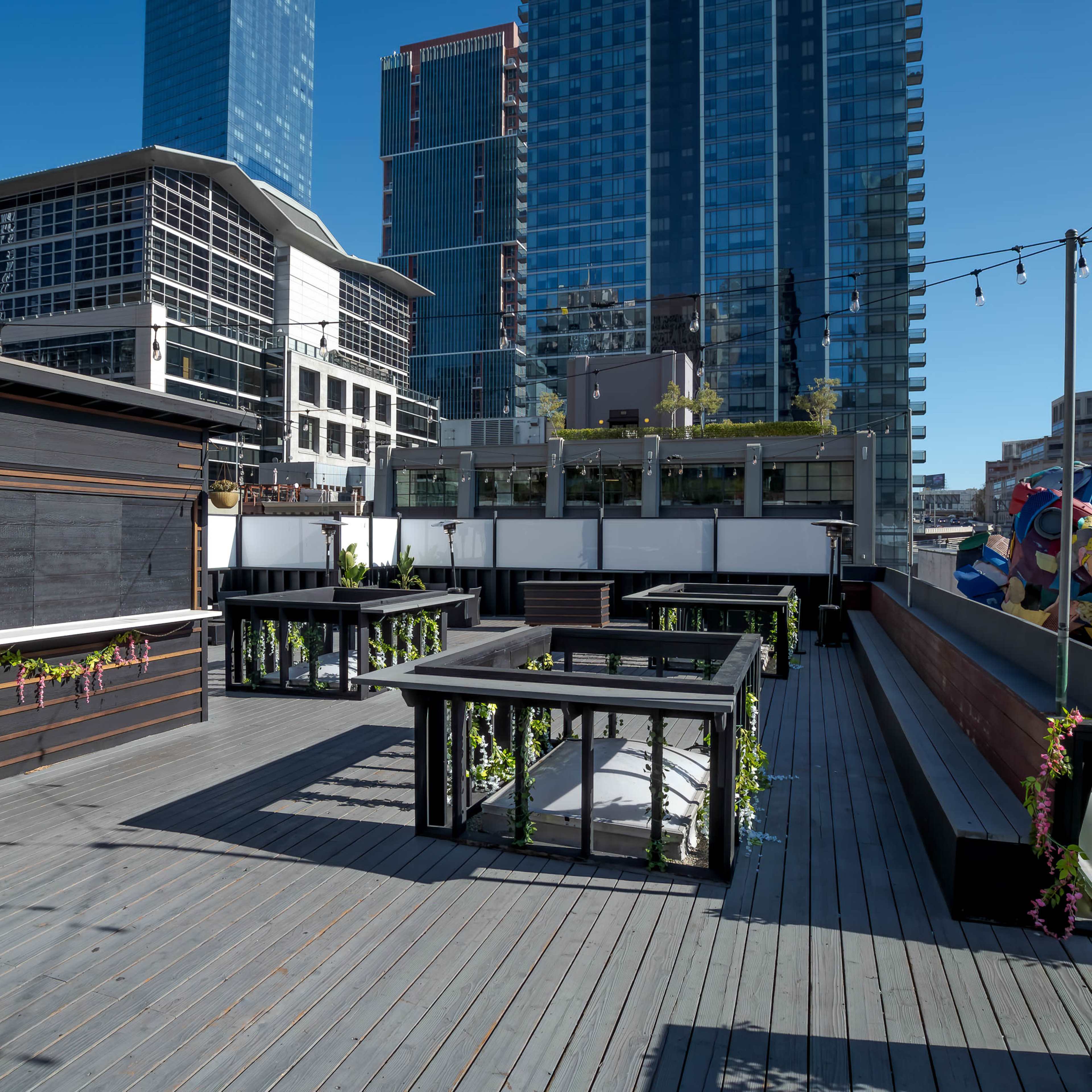 A rooftop area with wooden decking, garden planters, and structures set against a backdrop of modern high-rise buildings under a clear blue sky.