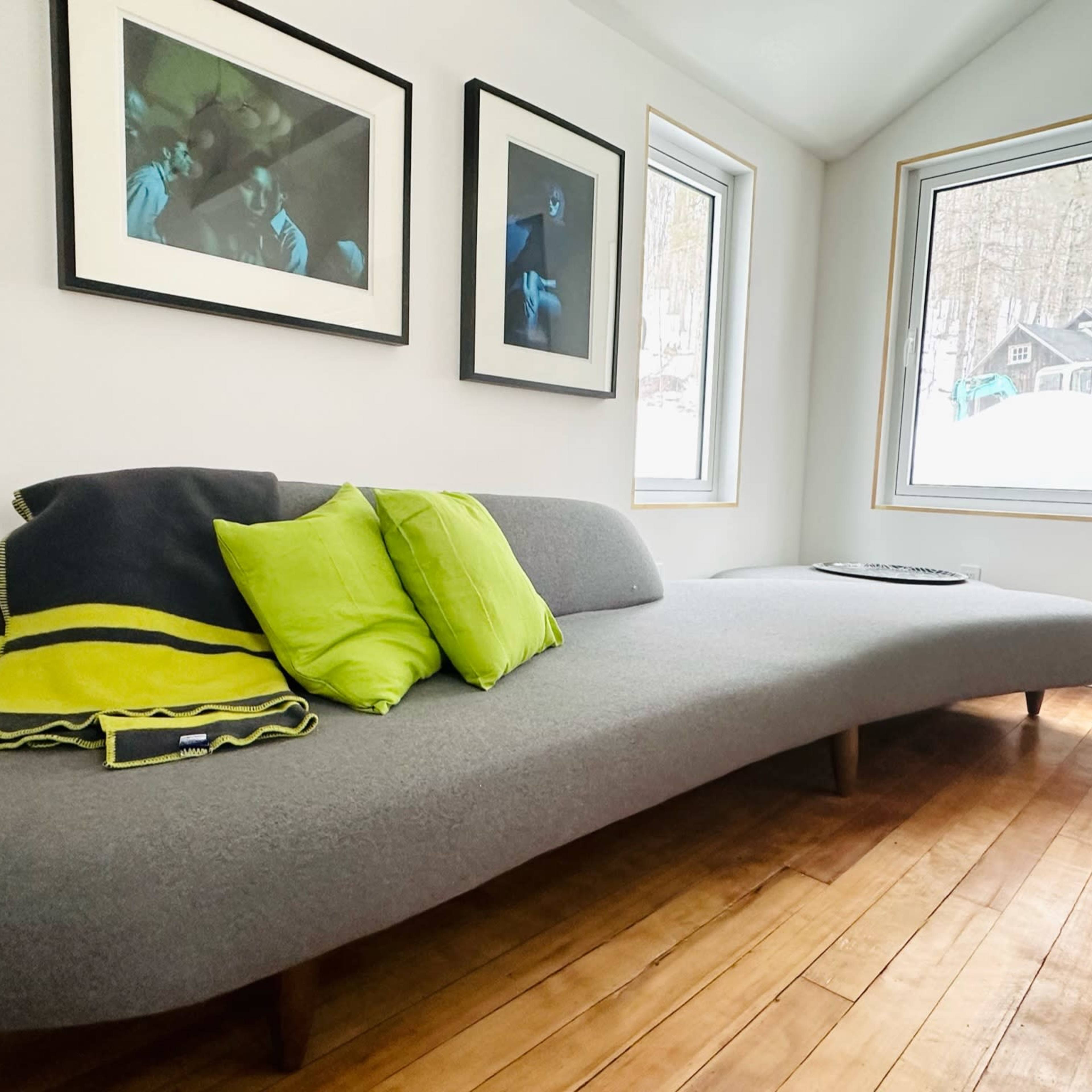 A modern, curved gray sofa with green accent pillows in a bright room featuring large windows and framed photographs on the walls.