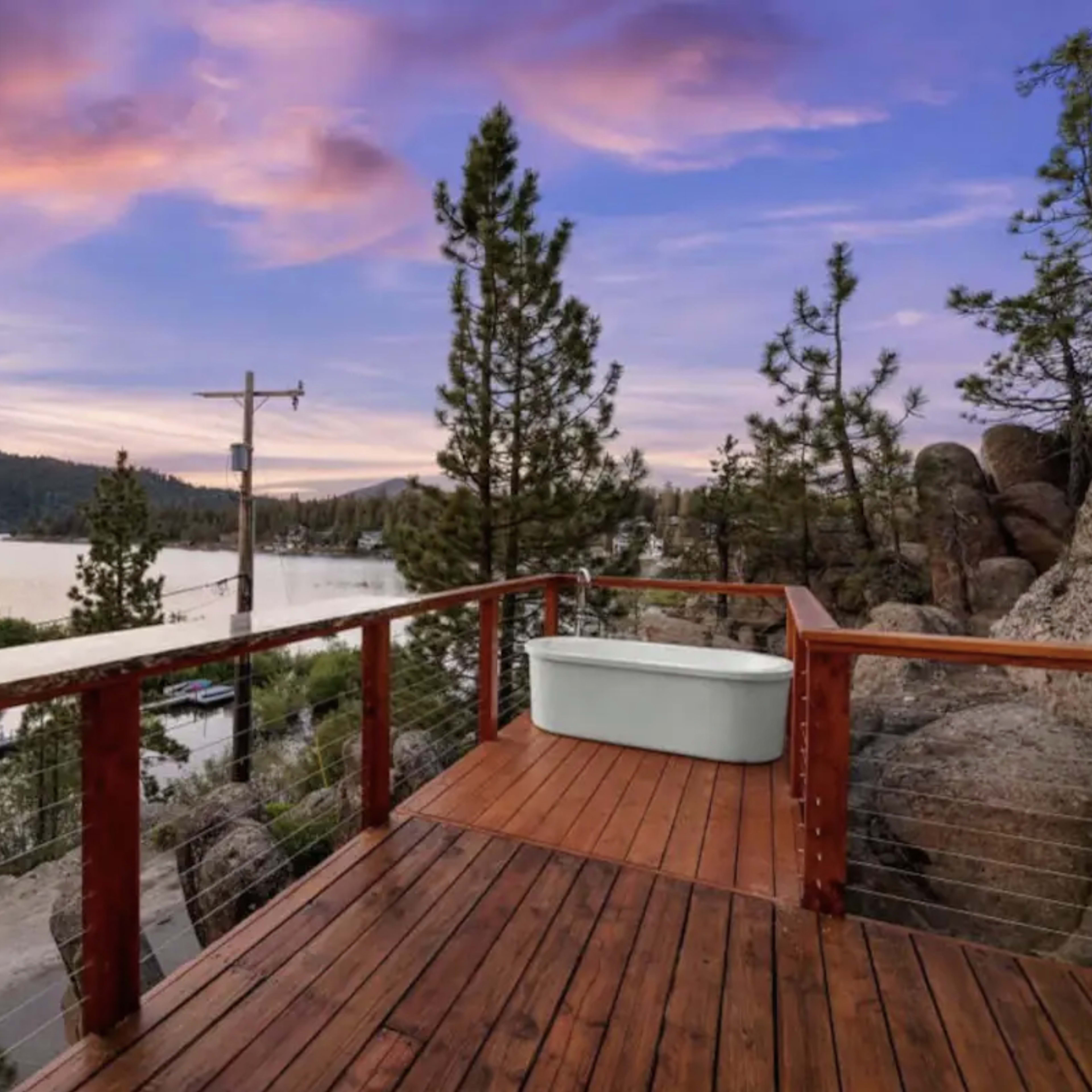 A wooden deck overlooks a tranquil lake surrounded by trees and rocky terrain under a colorful sky at dusk.