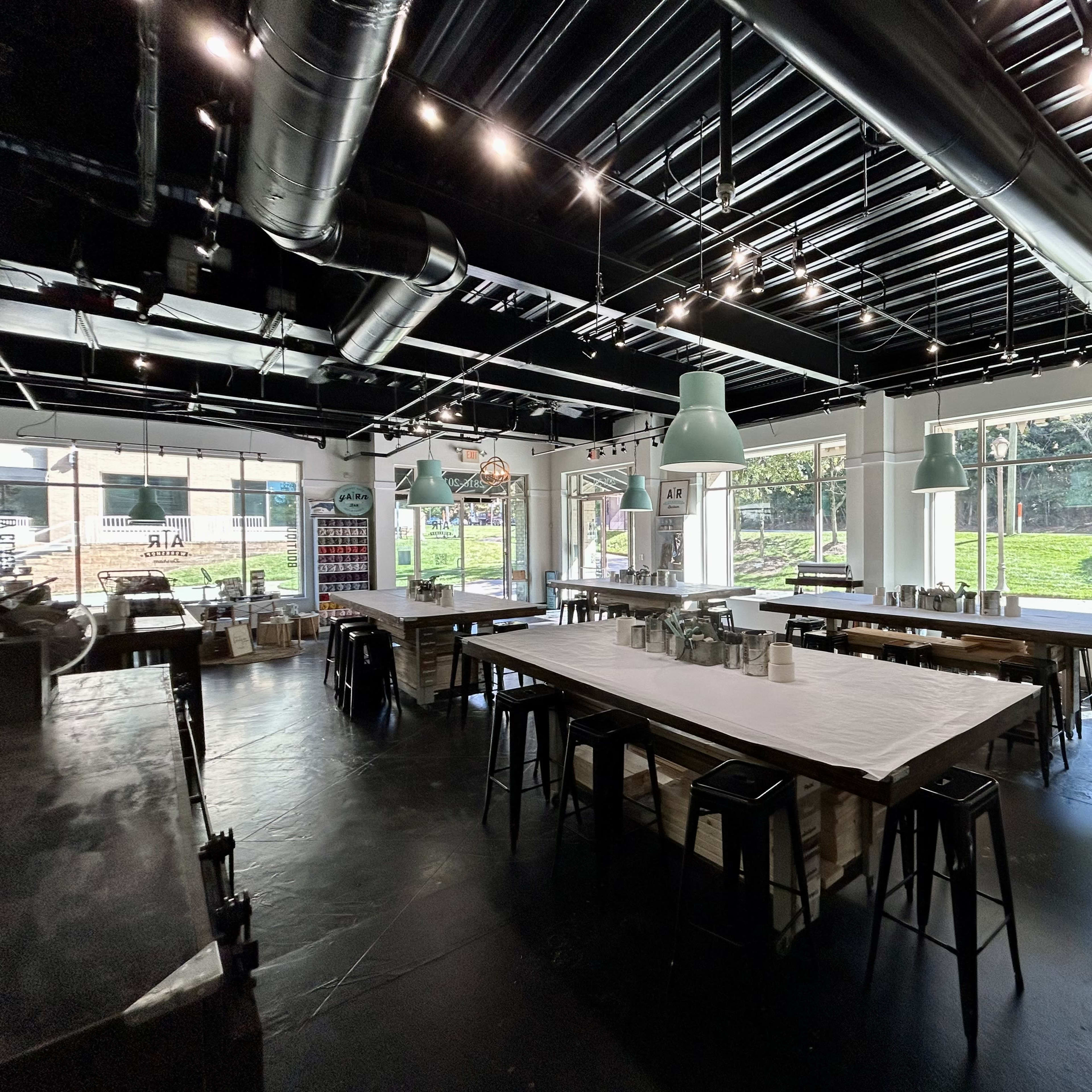 An industrial-style workspace with large tables, metal stools, and bright green pendant lights, illuminated by natural light from large windows overlooking an outdoor area.