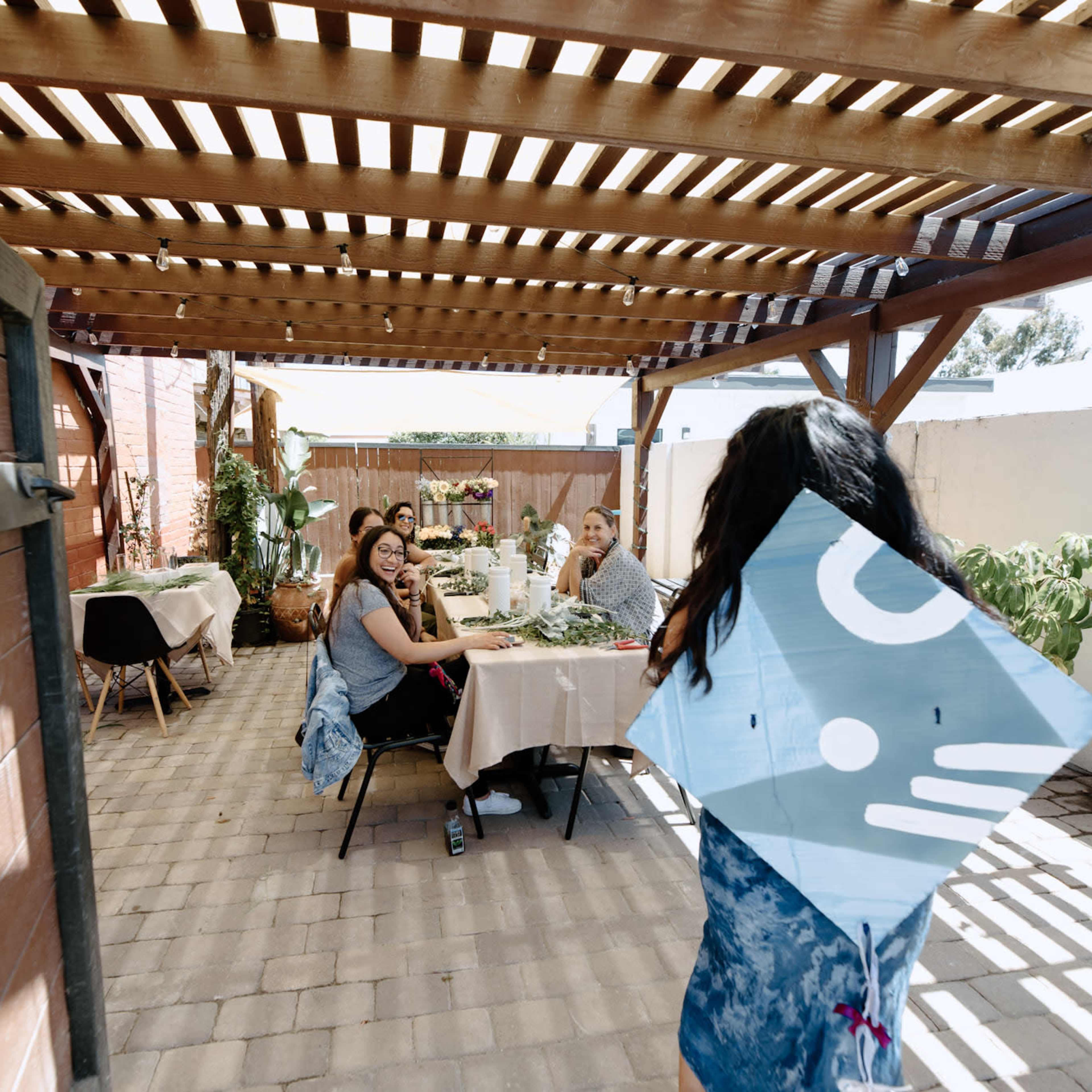 A group of people sits at a table under a wooden pergola while one person is walking towards them, holding a blue kite.