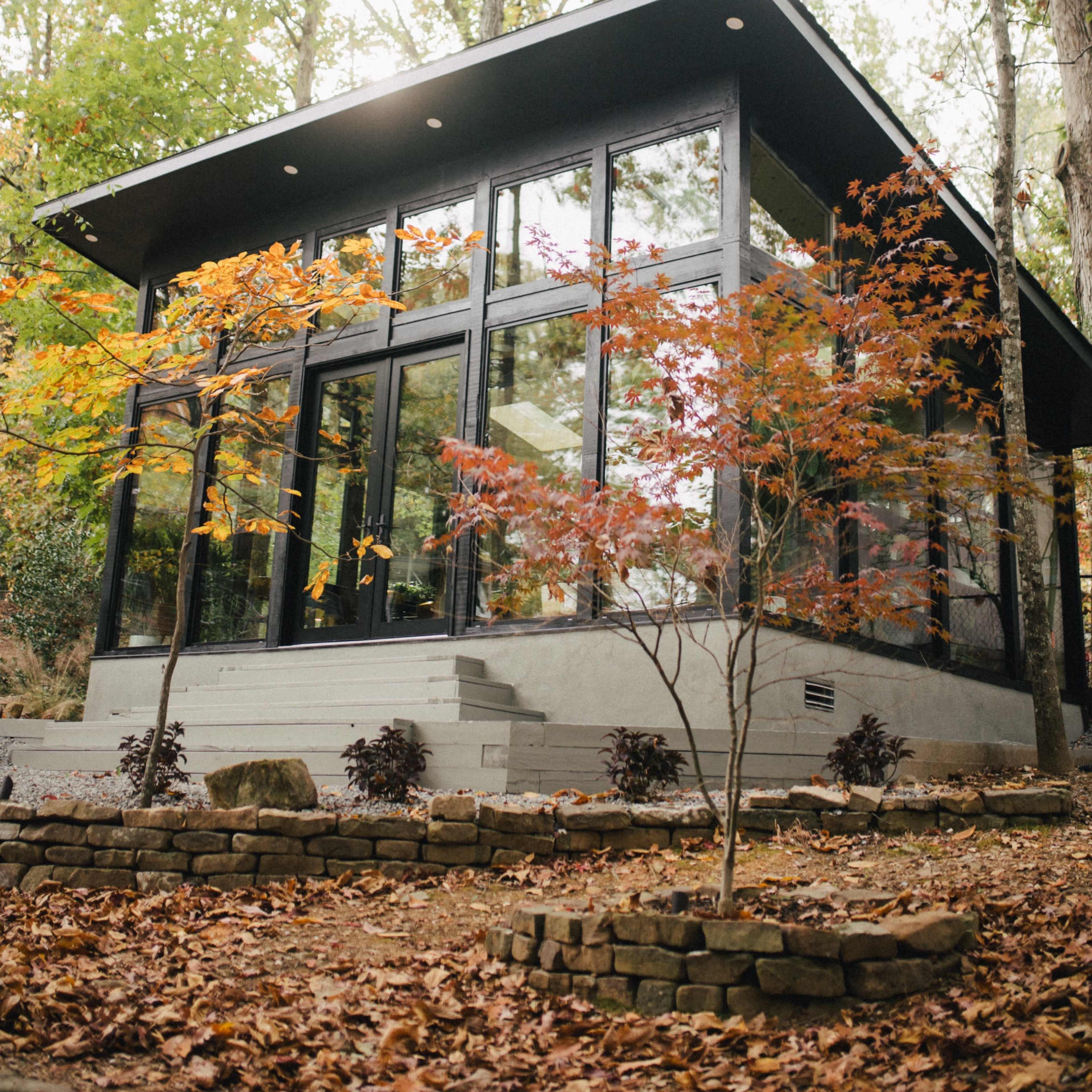 A modern, glass-fronted house is surrounded by trees with autumn foliage, set in a landscaped area with stone pathways.