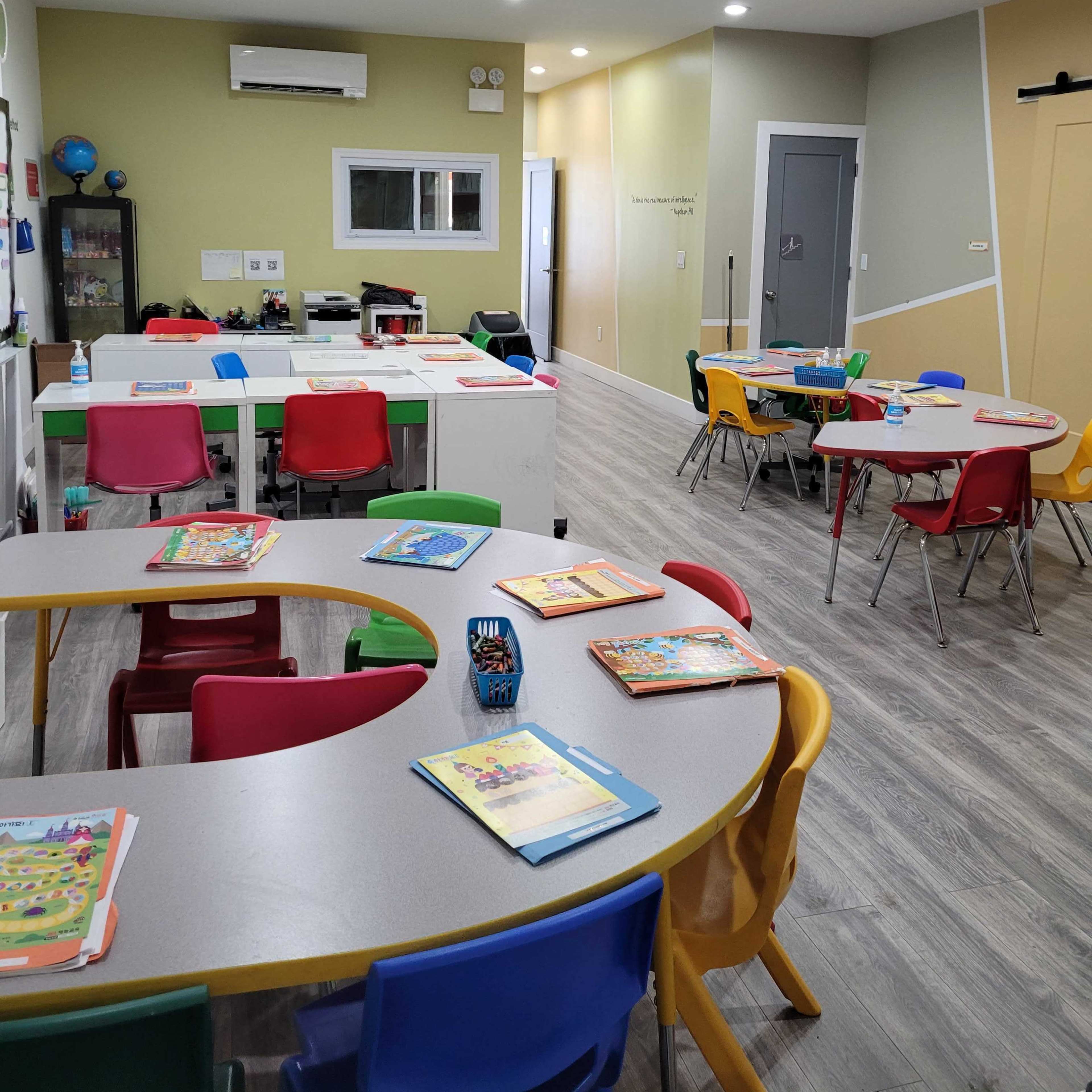 A brightly lit classroom features several tables with colorful chairs arranged, and books and materials laid out on the tables.
