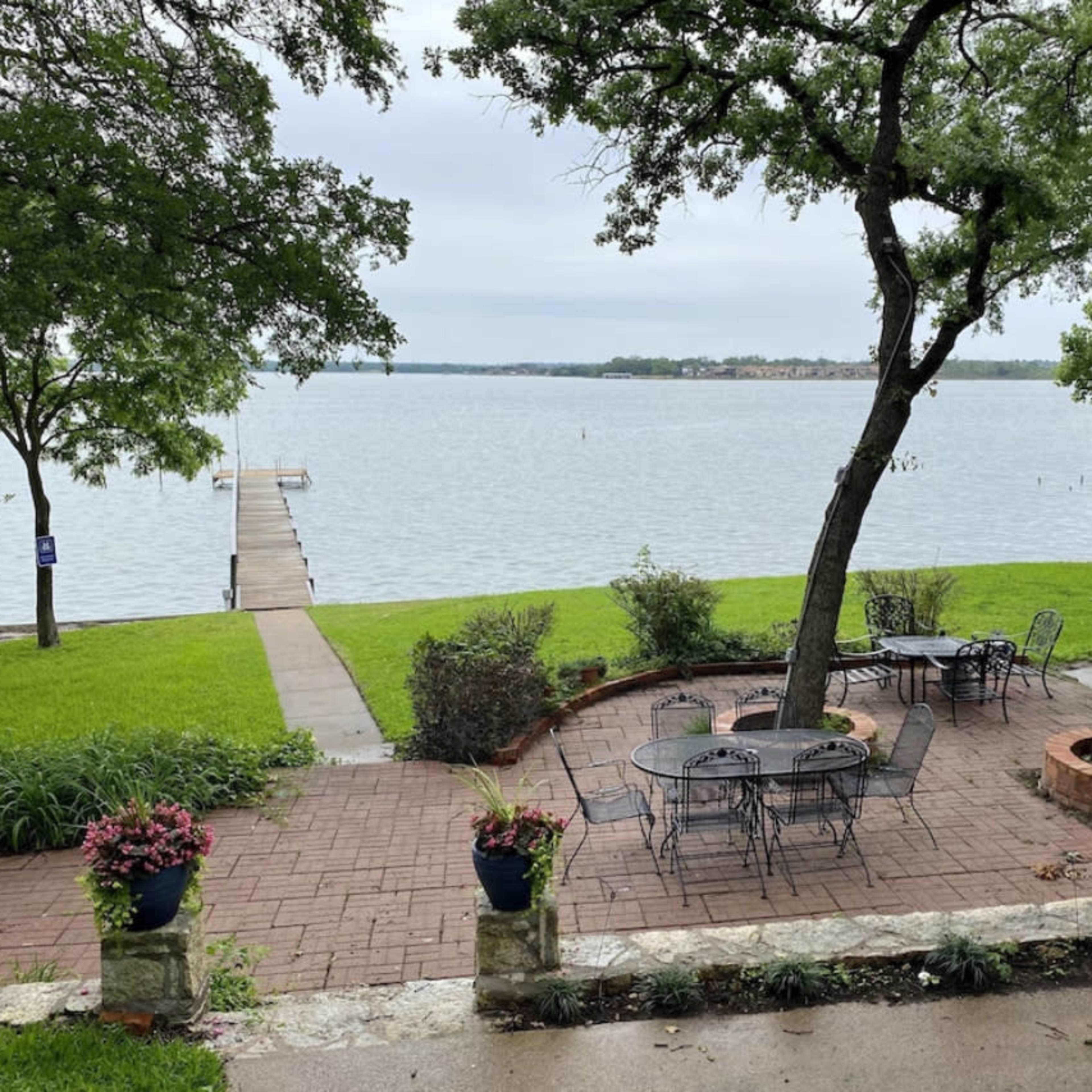 A patio with metal tables and chairs overlooks a calm lake, accompanied by a wooden dock extending into the water.