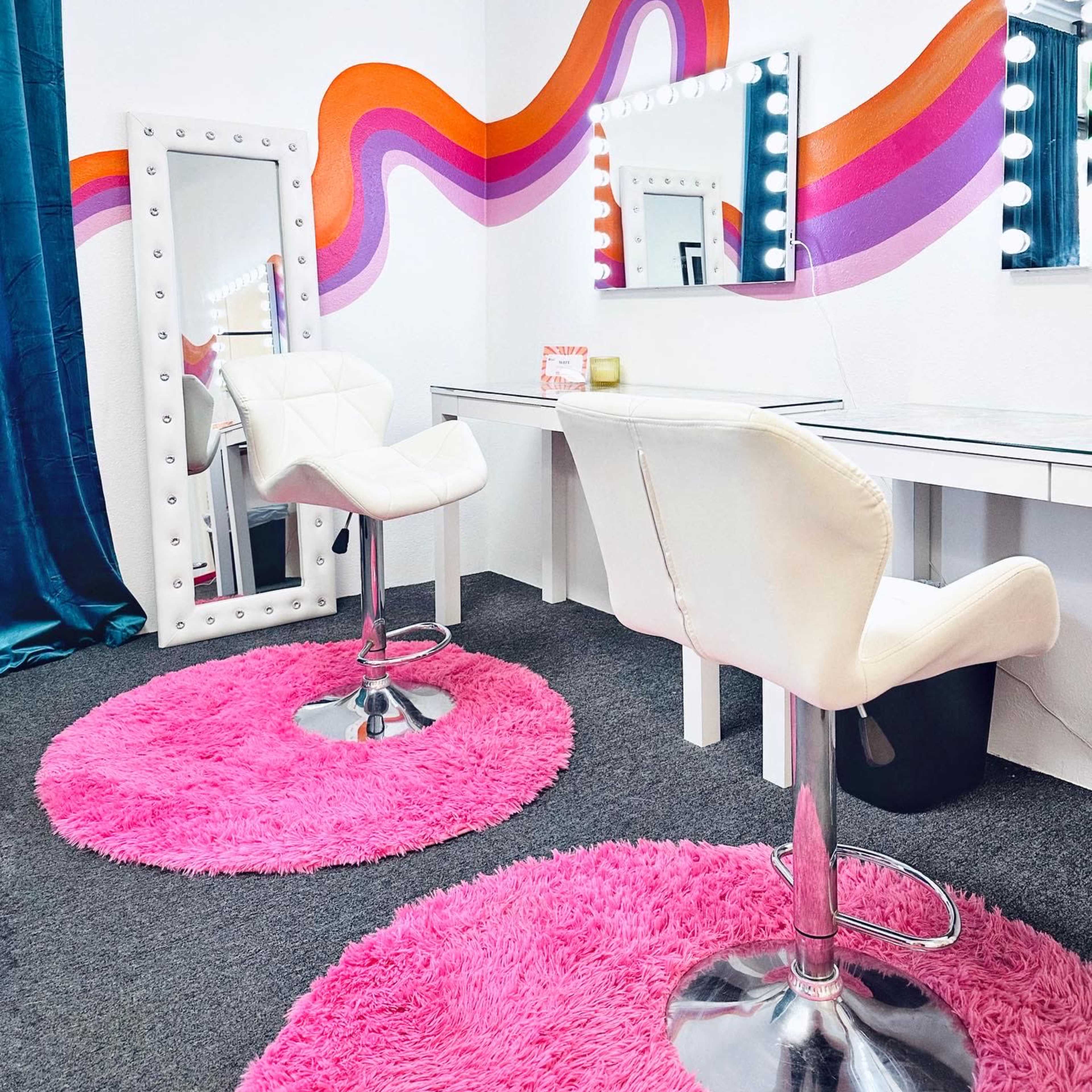 A brightly colored vanity area with two white bar stools, a large mirror framed with light bulbs, and a vibrant, wavy mural on the wall above.