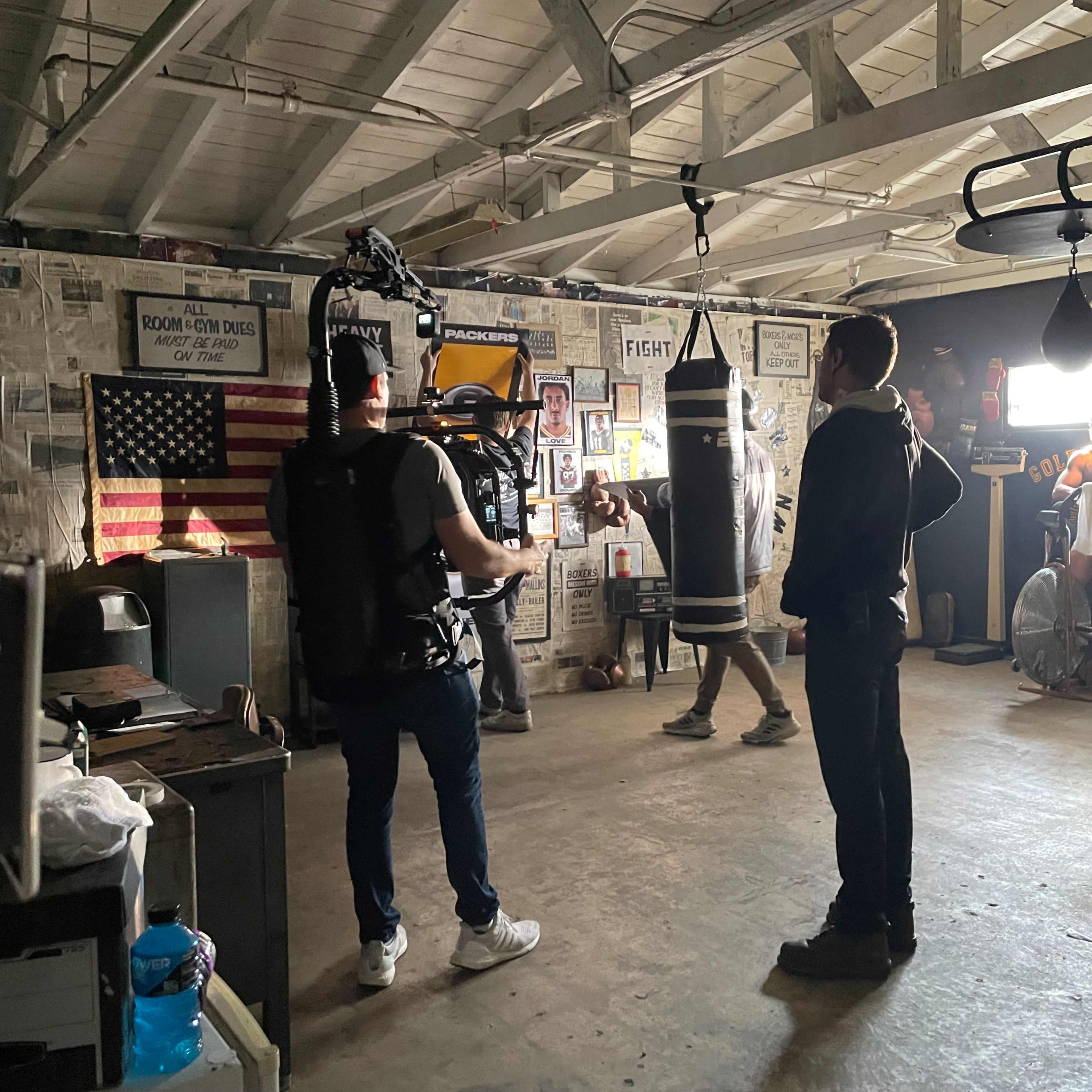 A film crew is shooting a scene in a boxing gym with a heavy bag and various boxing equipment visible on the walls.