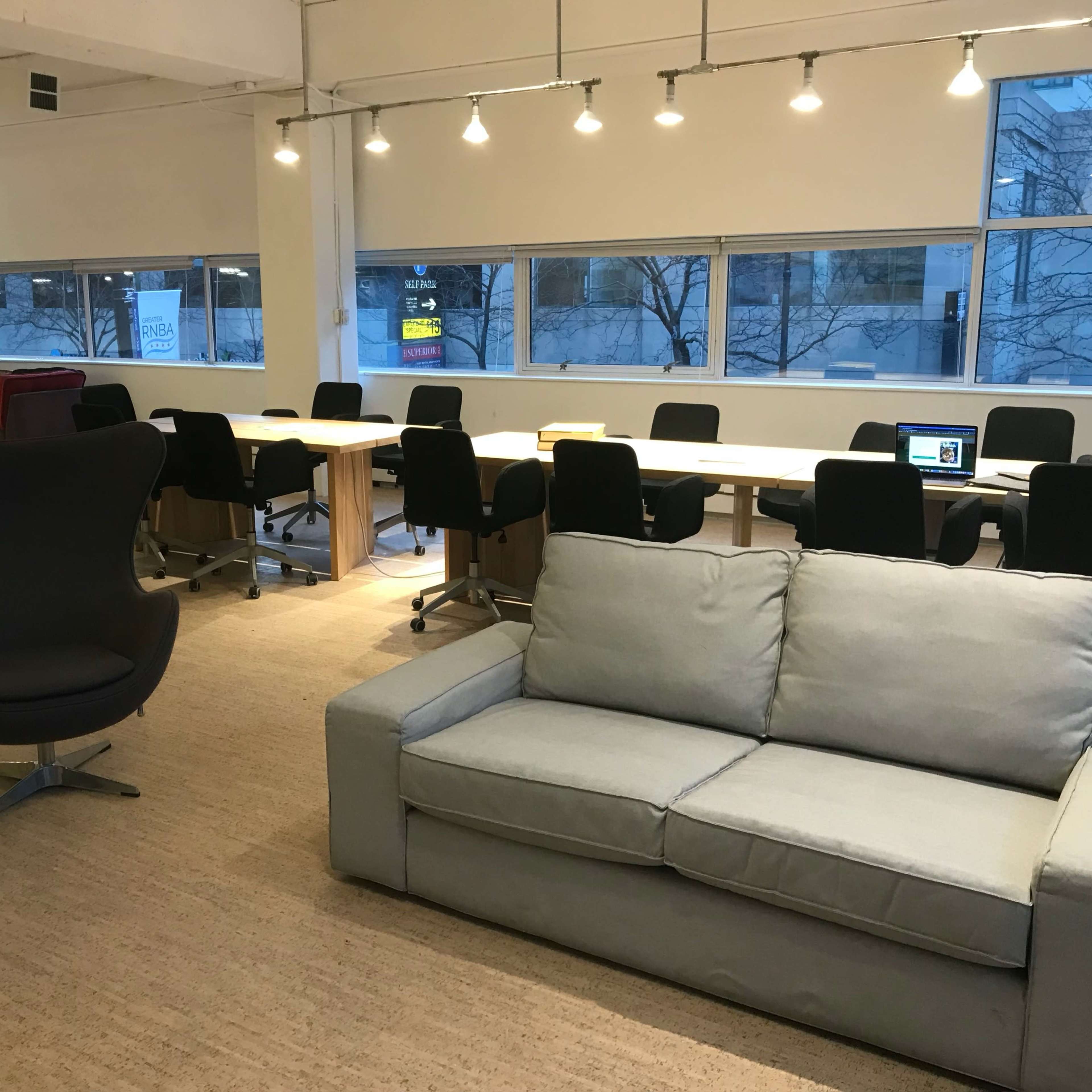 A spacious office area with a mix of seating arrangements, including a gray sofa, black chairs, and long tables beneath overhead lighting.