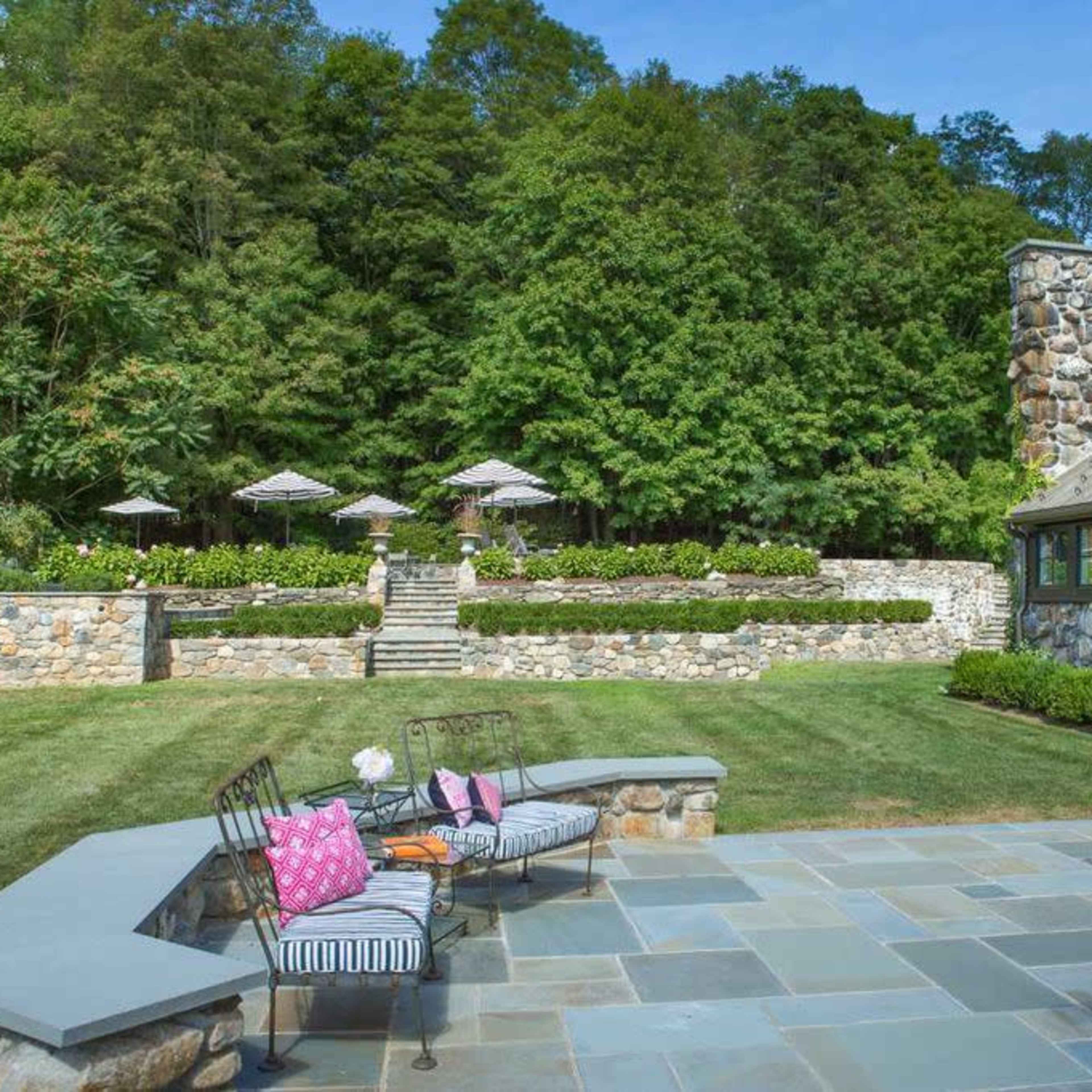 A landscaped backyard featuring a stone patio, a seating area with striped cushions, and steps leading to a garden with umbrellas and lush greenery in the background.