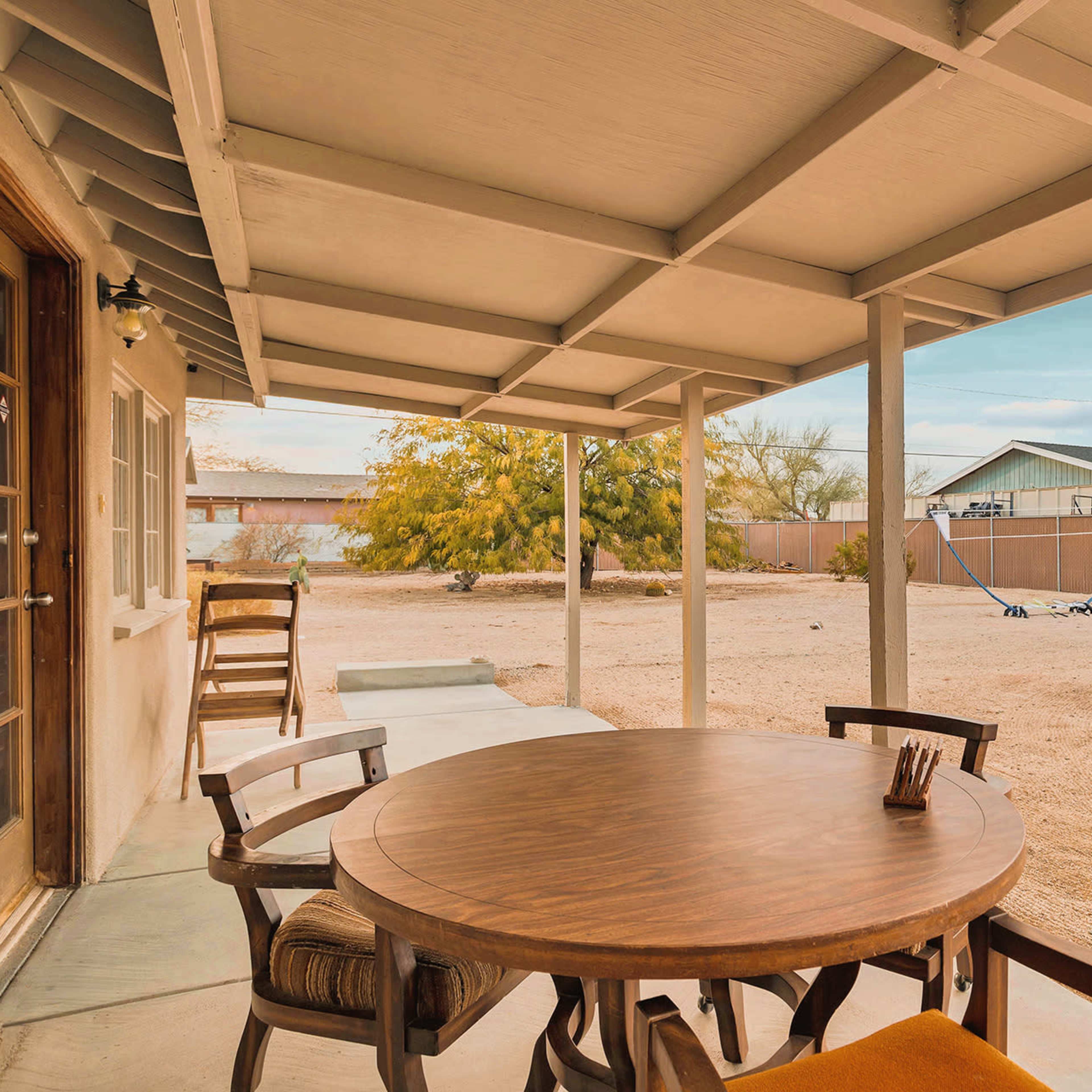 A wooden table with chairs is set under a covered patio, overlooking a sandy yard and neighboring structures in the background.