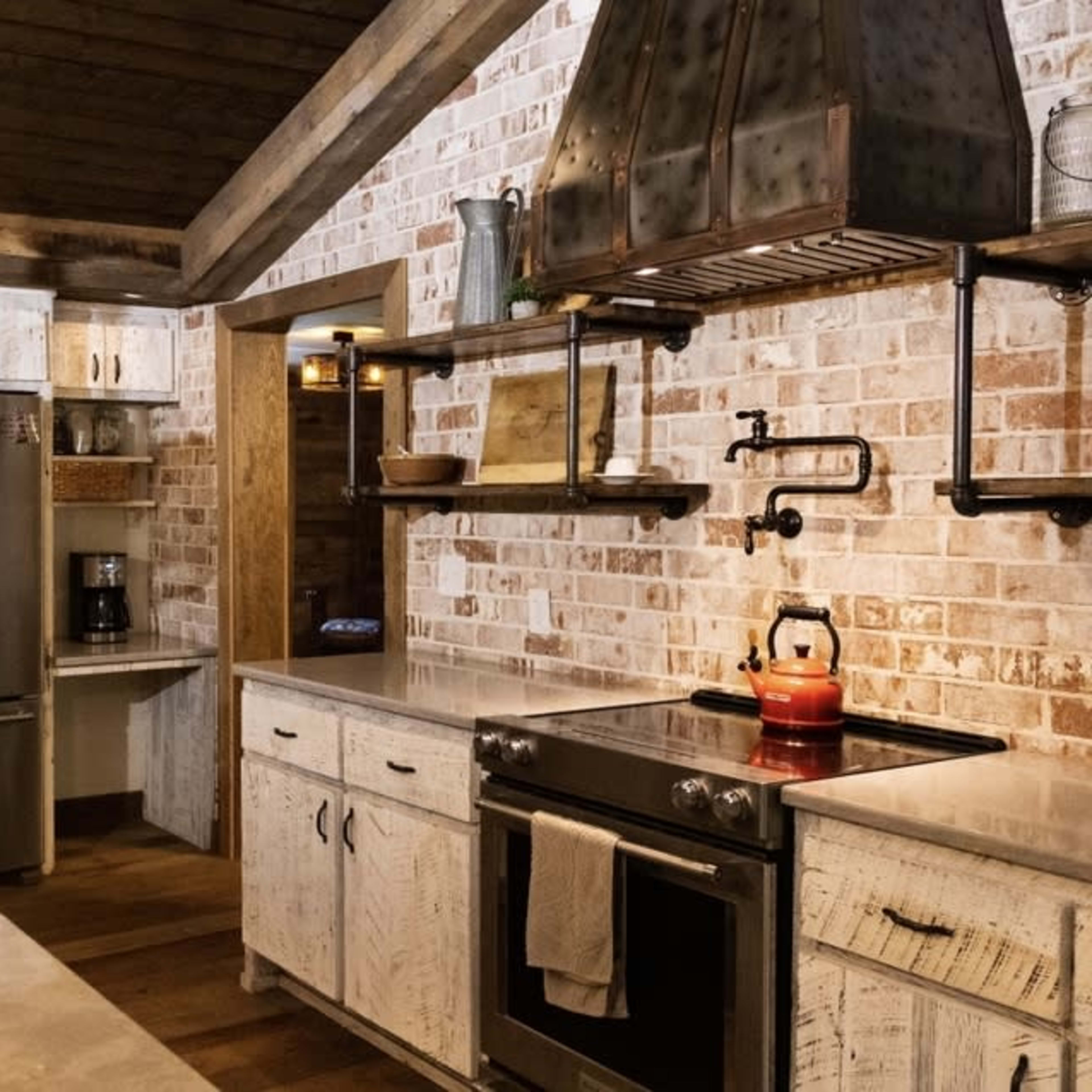 A modern kitchen with white wooden cabinetry, a stainless steel refrigerator, and a gas stove under a metal hood, featuring exposed brick walls and wooden beams.