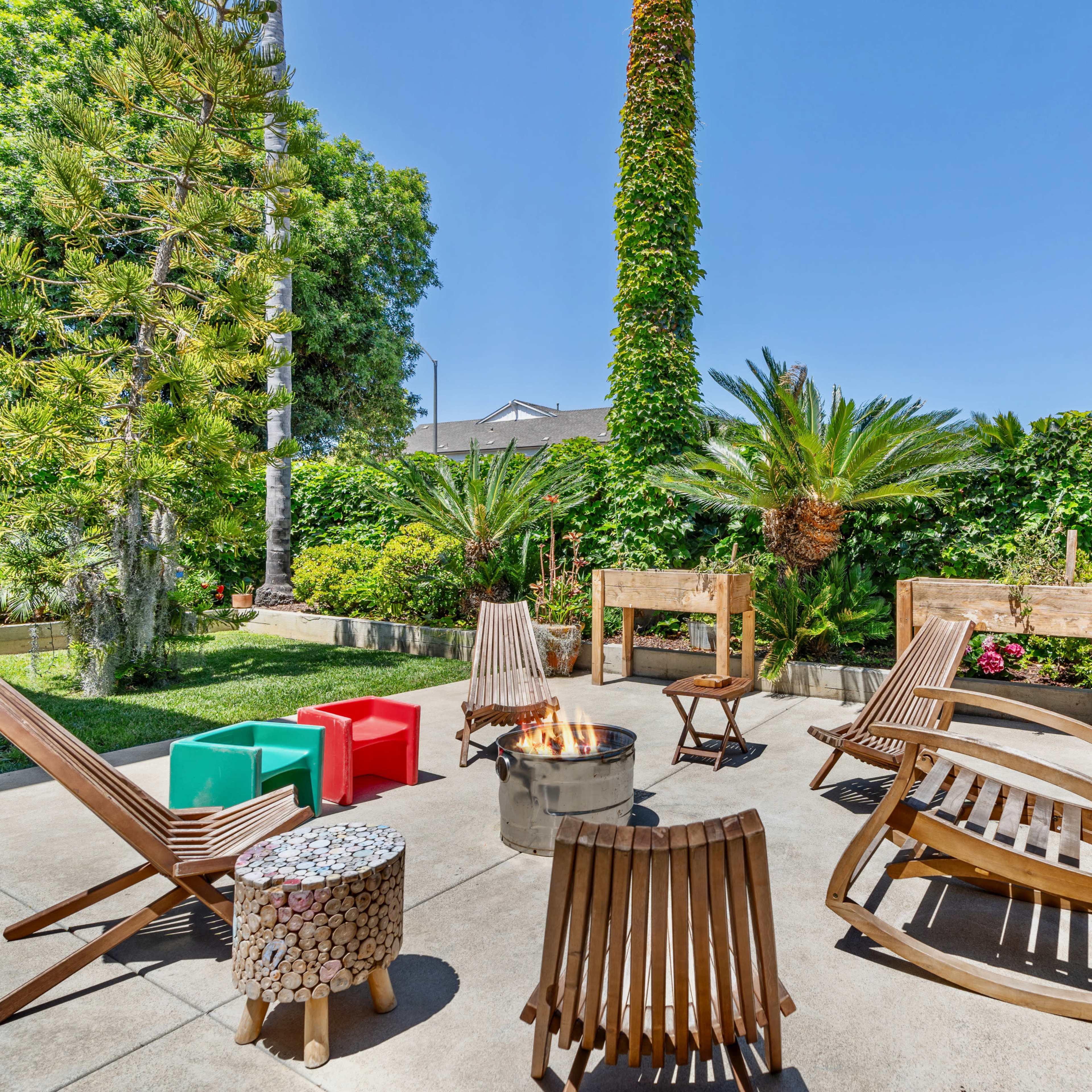 A patio area features wooden chairs and a fire pit surrounded by lush greenery and tall palm trees.