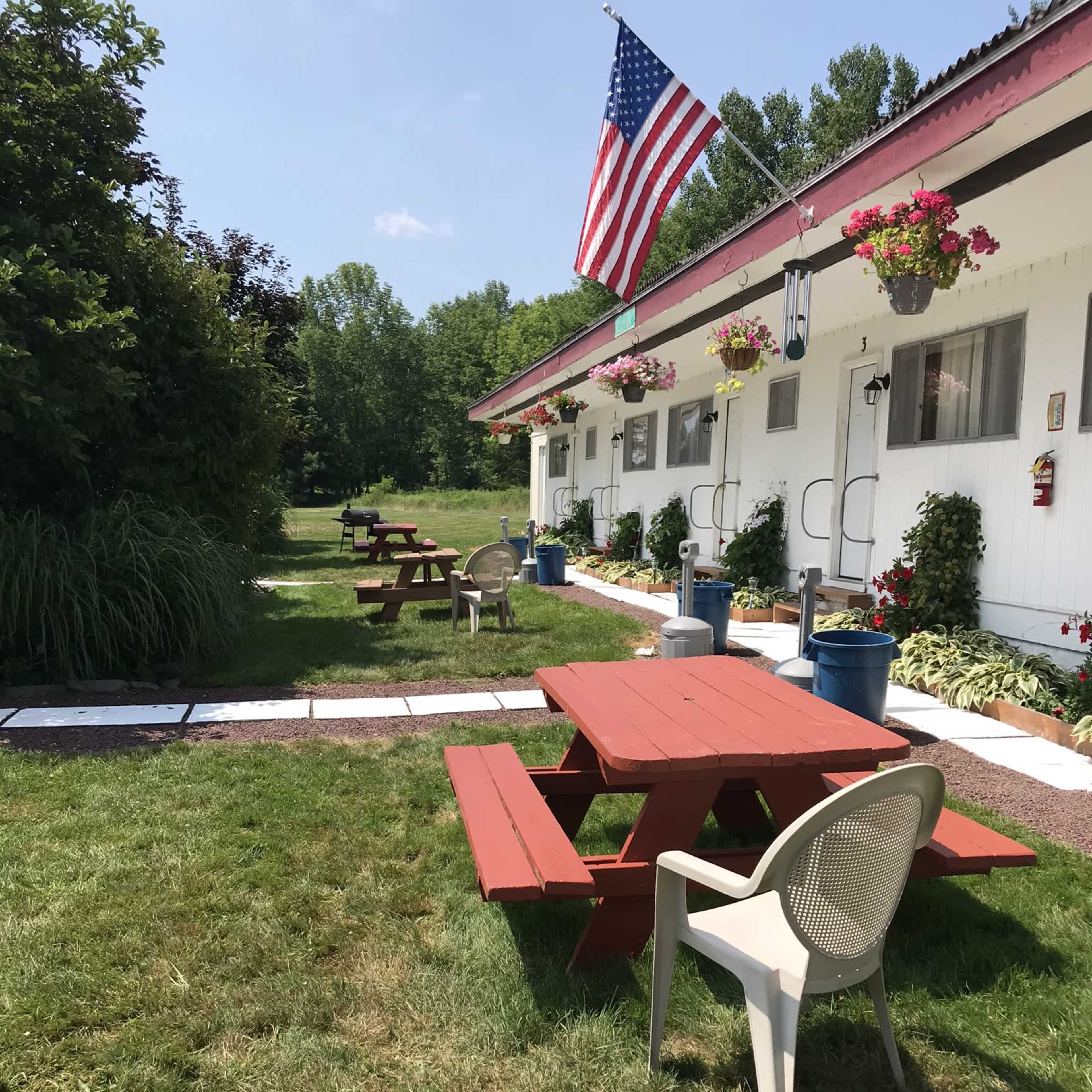 A grassy area with red picnic tables and a white building, featuring an American flag and flower pots along the exterior.