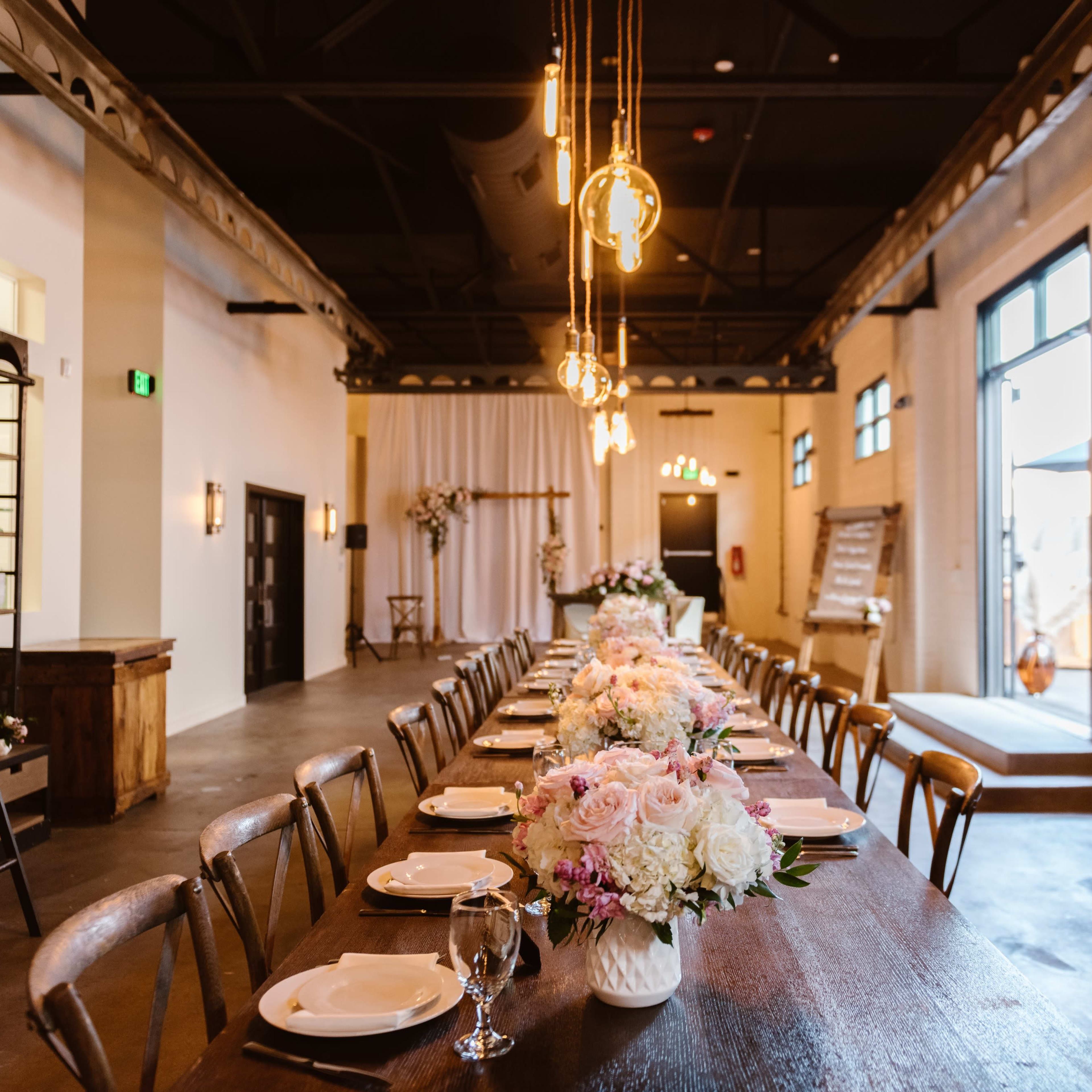 A long dining table is set with plates and floral arrangements in a spacious, well-lit venue.