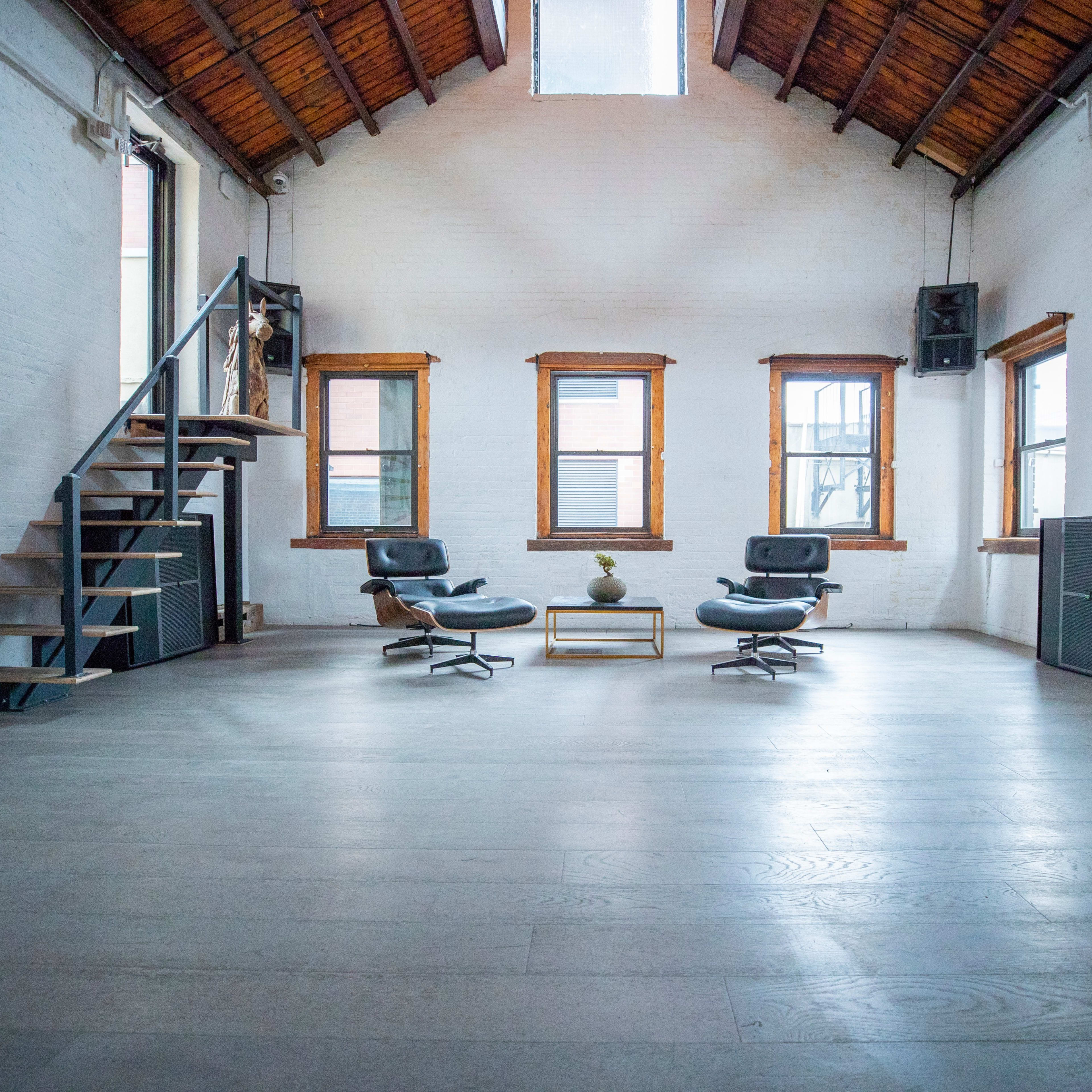 A spacious room with wooden beams, featuring two black chairs, a small table, and a staircase leading to a second level.
