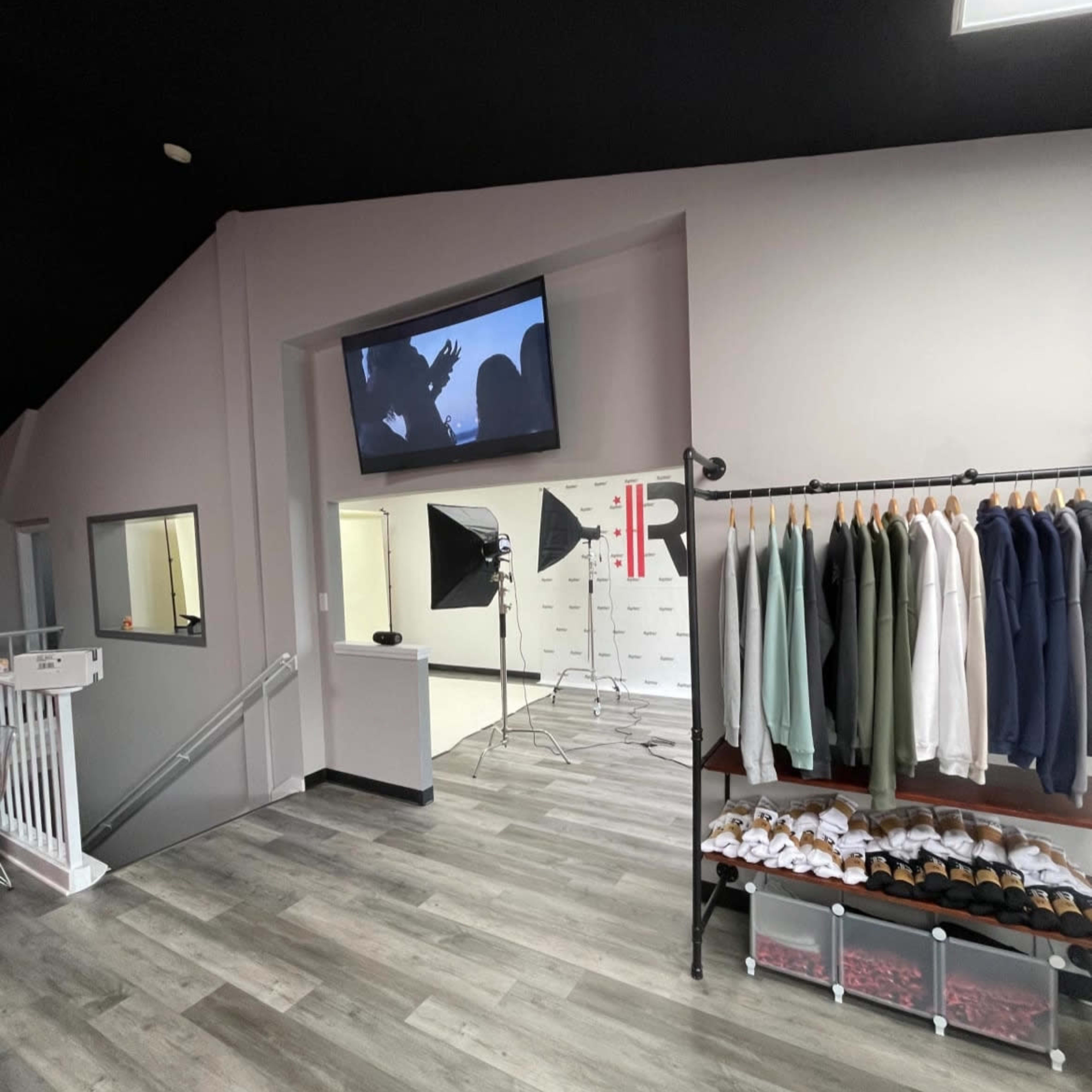 A modern retail space with gray walls, wood flooring, organized clothing racks, and a TV displaying a video in the background.