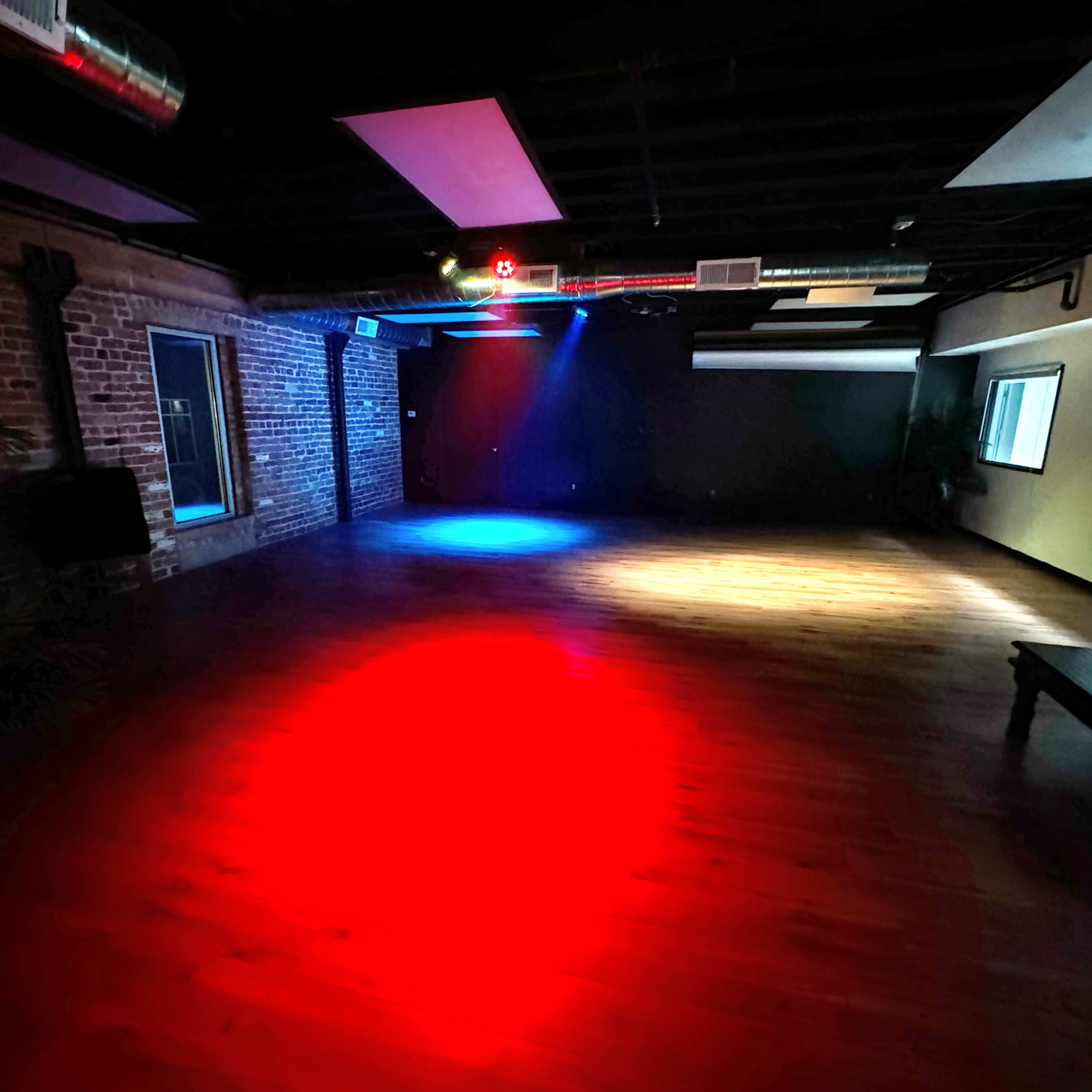 The room features hardwood flooring illuminated by red and blue lights, with exposed brick walls and several windows.