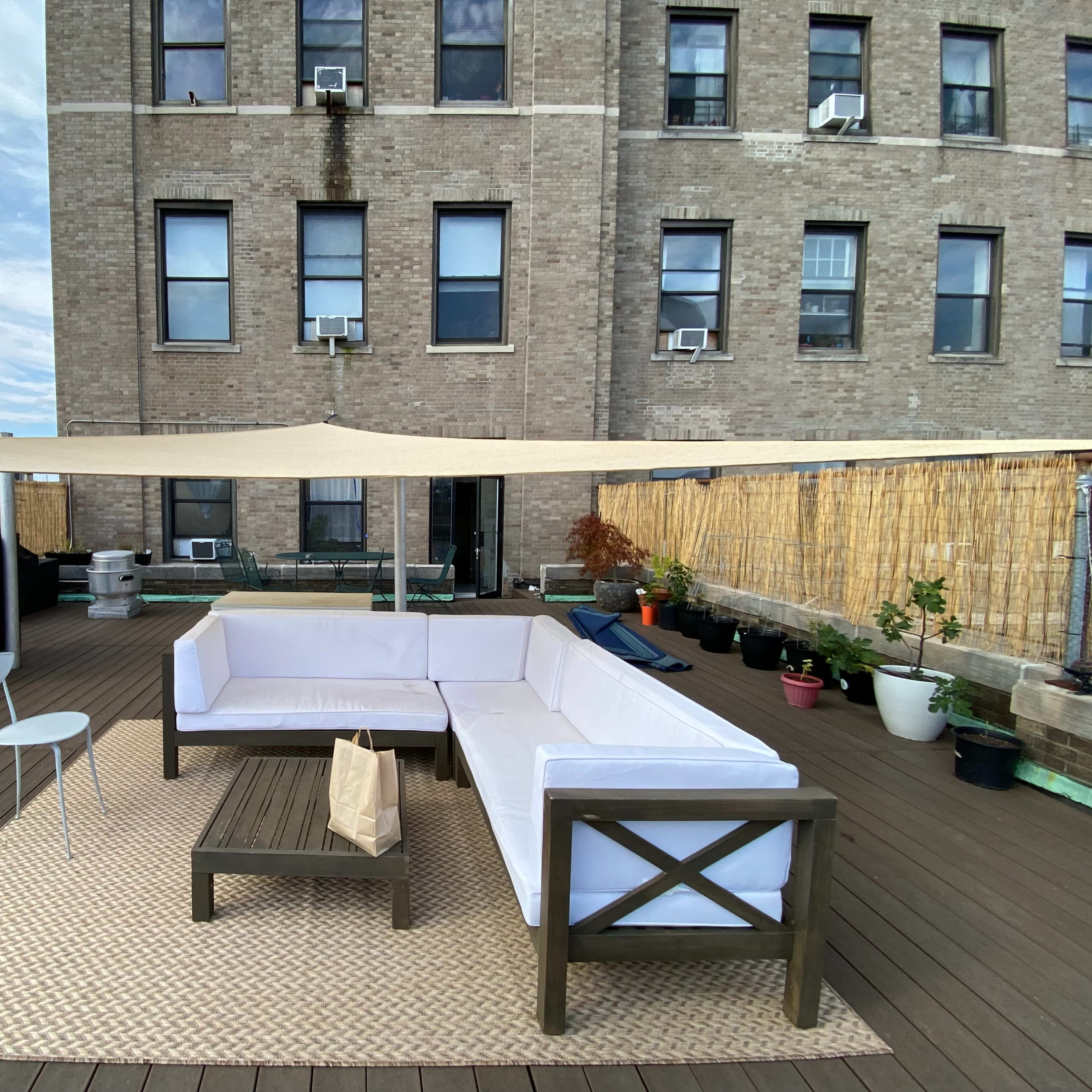 A rooftop patio features a sectional sofa, a coffee table, and potted plants under a canopy, with an apartment building in the background.